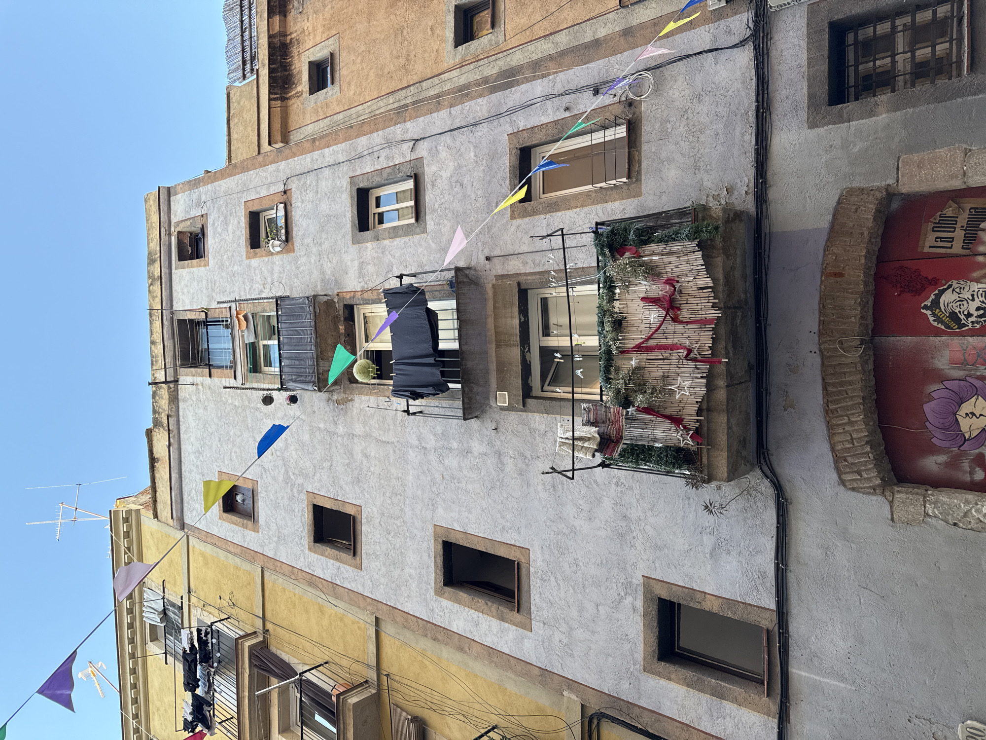 This is a low-angle photograph of the exterior of an old building in Barcelona, Spain. The building is multi-story with a mix of yellow and gray textured walls, and a variety of window types. Festive triangular flags are strung across the facade, adding color. Balconies, some with decorative coverings, extend from some windows. The building has exposed electrical wiring running along its side, and graffiti on the wall next to a red door on the right. Clothes hang from a clothesline outside a window. The sky is blue.