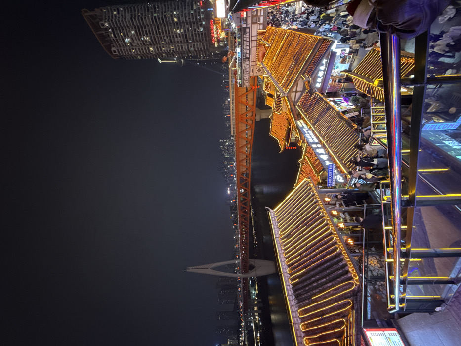 The image presents a panoramic nighttime view of Hongyadong, located in Yuzhong District, Chongqing, China. The foreground is dominated by a multi-level complex of traditional Chinese-style stilt houses (diaojiaolou), distinguished by dark tiled roofs and facades extensively outlined with warm-toned LED strip lights. Modern glass and metal railings of a viewing platform or walkway are prominent in the immediate lower foreground, with visible escalators facilitating pedestrian movement between levels. A dense crowd of people populates the various tiers of the traditional complex, moving through what appear to be commercial establishments. Beyond these structures, the dark surface of the Jialing River reflects scattered city lights. Spanning the river are components of the illuminated Qianximen Bridge, including a prominent orange truss section and a white cable-stayed pylon. The background features a dense urban skyline of numerous illuminated modern high-rise buildings on both riverbanks, with a particularly tall, curvilinear skyscraper visible on the far right. The sky is uniformly dark, indicating a nighttime setting.