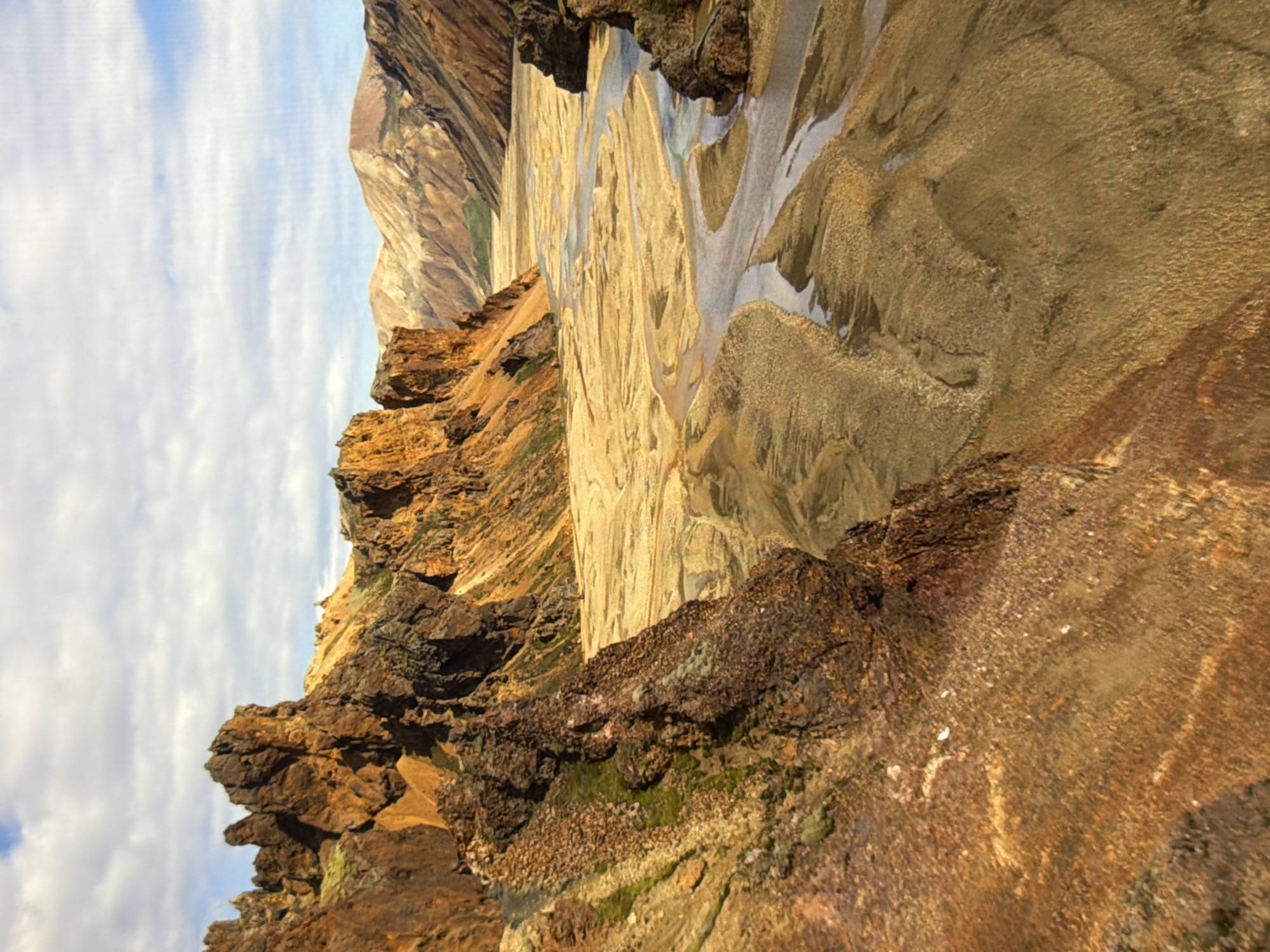 This image depicts a vast, arid landscape with rugged, colorful mountains and a braided river system. The mountains are predominantly ochre and brown, with some patches of green vegetation clinging to their slopes. The river carves its way through a sandy plain, with multiple channels converging and diverging. The sky is partly cloudy, with white clouds scattered across a blue expanse. The scene conveys a sense of natural grandeur and wilderness. The image is not located in the city of Kyiv, Ukraine; it is a natural landscape.