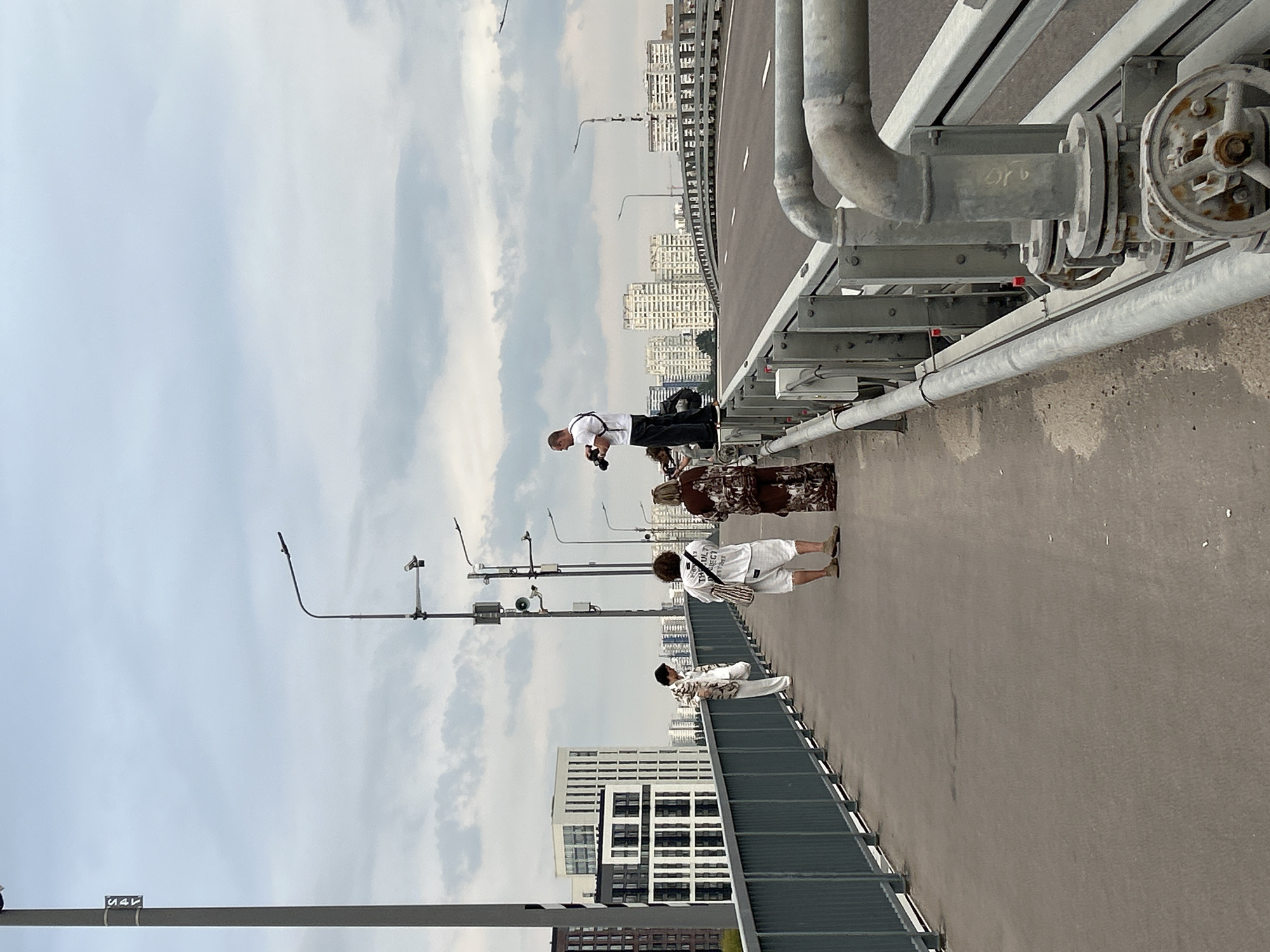 A group of four people are on a pedestrian walkway of a bridge over a road. The man furthest to the right is standing on the railing, holding a camera and taking a picture. Next to him, to his left, is a woman in a dark patterned dress. In front of her are two more people. One is a person in white shorts and a white t-shirt with a bag over their shoulder. The other person is in white pants and a patterned shirt, facing away from the camera. In the background are buildings and a cloudy sky. There are streetlights and pipes visible on the bridge.