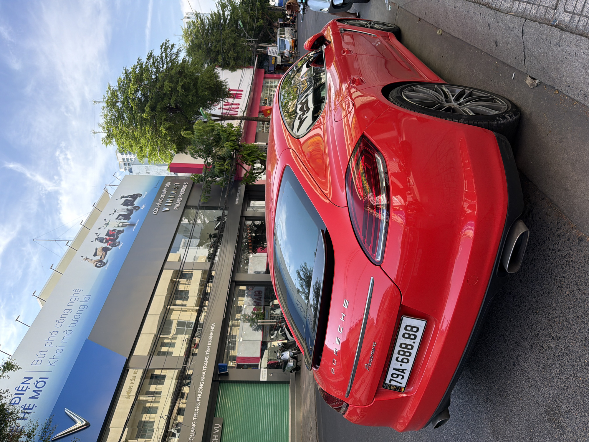 A bright red Porsche Panamera is parked on the side of a city street in Nha Trang, Vietnam. The car is positioned at an angle, facing away from the camera and slightly to the left. Its license plate reads "79A-688.88". The car is a prominent subject, occupying the foreground and center of the image. Behind the car, buildings line the street, featuring shopfronts with green metal shutters and signage. One sign, in Vietnamese, appears to be for an electric vehicle dealership, displaying images of scooters. The street is paved, and a concrete sidewalk is visible. Trees with lush green foliage are interspersed among the buildings, and a blue sky with some white clouds is overhead. The overall scene depicts a typical urban streetscape in Vietnam.