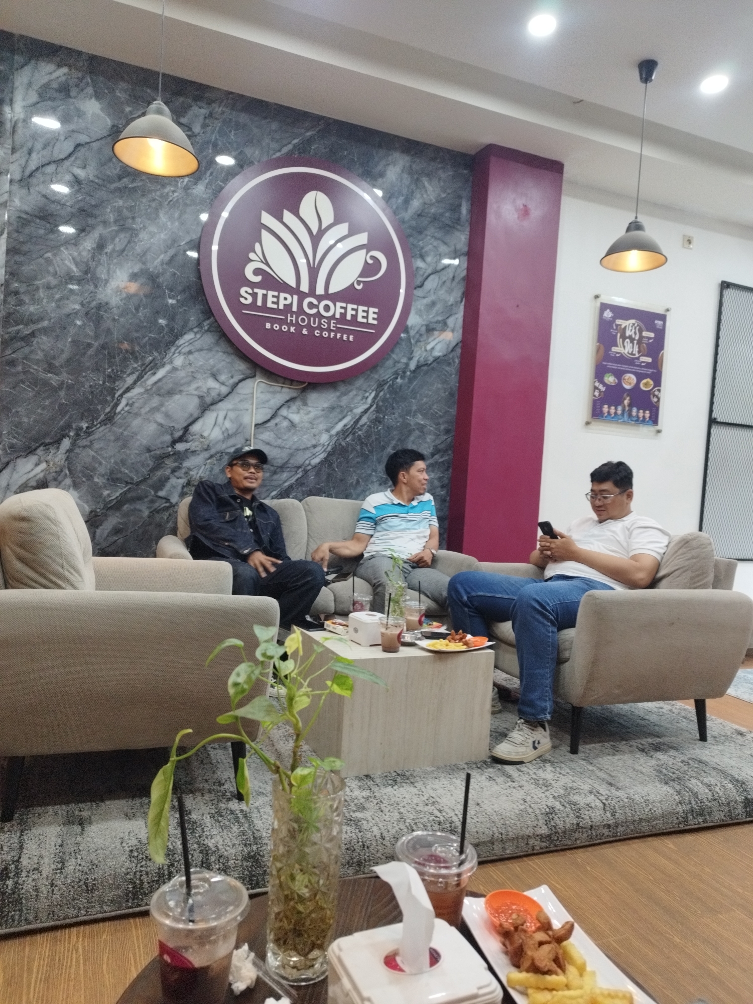 In a South Jakarta coffee shop, three men are seated in a lounge area. The man on the left, wearing a dark denim jacket and a baseball cap, sits on a cream-colored sofa, looking to his right. In the center, another man in a blue and white striped shirt is seated on the same sofa, facing towards the right side of the frame. To the right, a third man in a white polo shirt and blue jeans is seated on a separate cream-colored armchair, looking down at his mobile phone. A low, light-colored coffee table sits between the sofas, with drinks and a plate of food, including fries and what appears to be chicken nuggets, on it. A decorative plant in a glass vase is also on the table. The wall behind them features a dark marble-like pattern with a circular logo for "STEPI COFFEE HOUSE - BOOK & COFFEE." Two pendant lights hang from the ceiling, illuminating the space. The floor is covered with a grey patterned rug over wooden flooring. The overall ambiance suggests a casual gathering at a coffee shop.