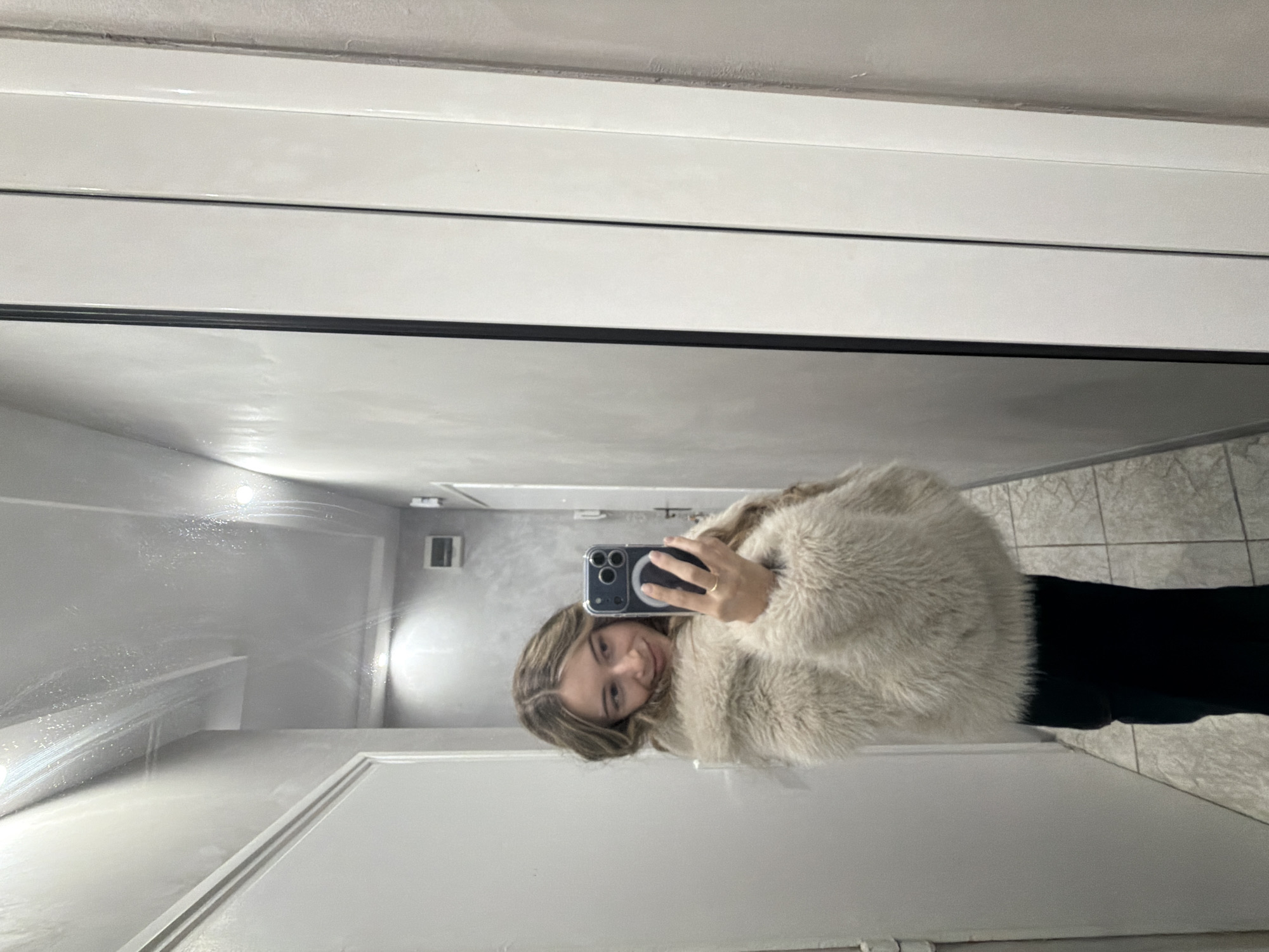 A woman takes a mirror selfie in what appears to be a bathroom. The walls are white, with tiling visible in the reflection suggesting a bathroom setting. She is wearing a fluffy cream-colored jacket and dark trousers. The phone she's using is visible, held in her right hand. The location isn't discernible as Plovdiv, Bulgaria, based on the visual details of the image.
