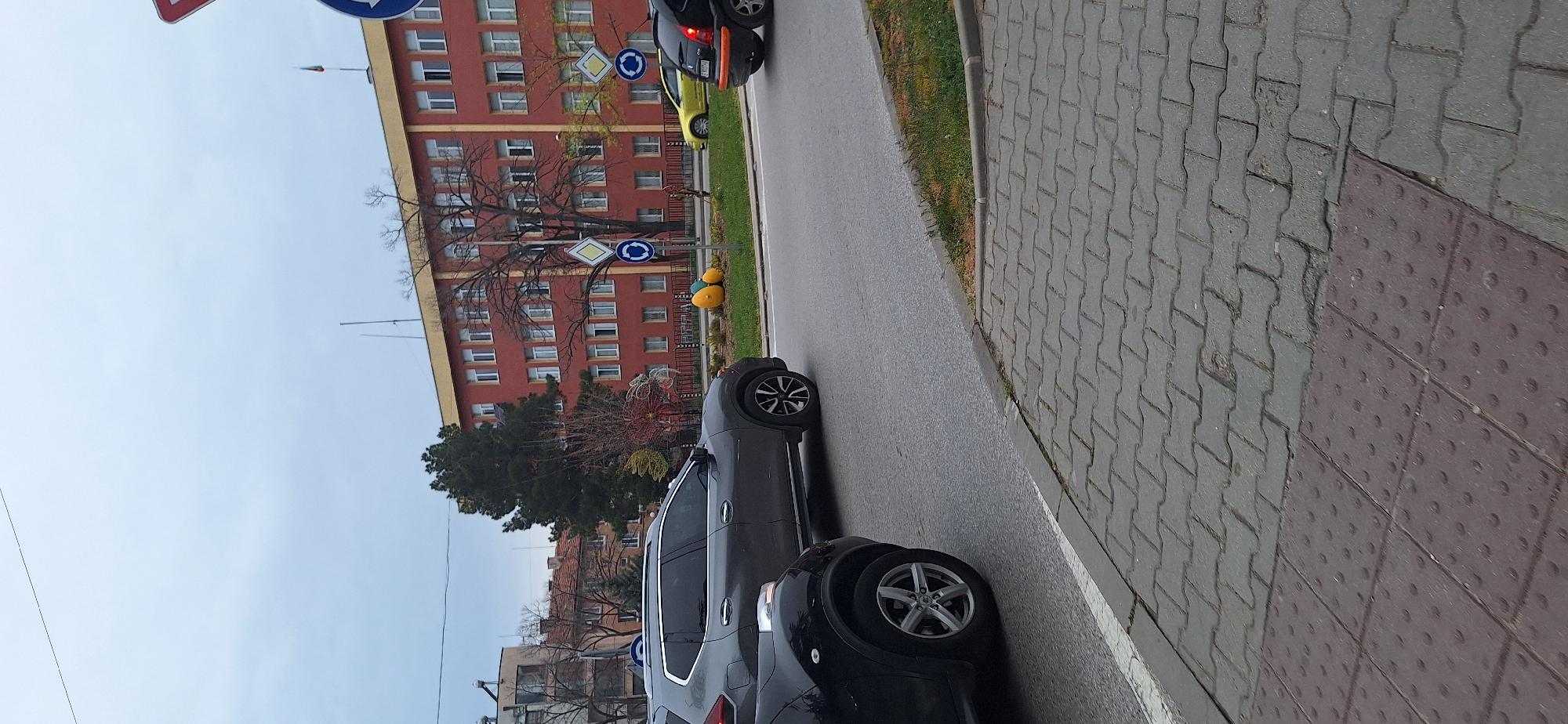 The image shows a street scene in Blagoevgrad, Bulgaria. Several cars are parked on the side of the road, including a gray SUV and a black compact car. Street signs are visible near a multi-story red brick building with numerous windows. A sidewalk paved with gray pavers is on the right side of the frame. The sky is overcast.
