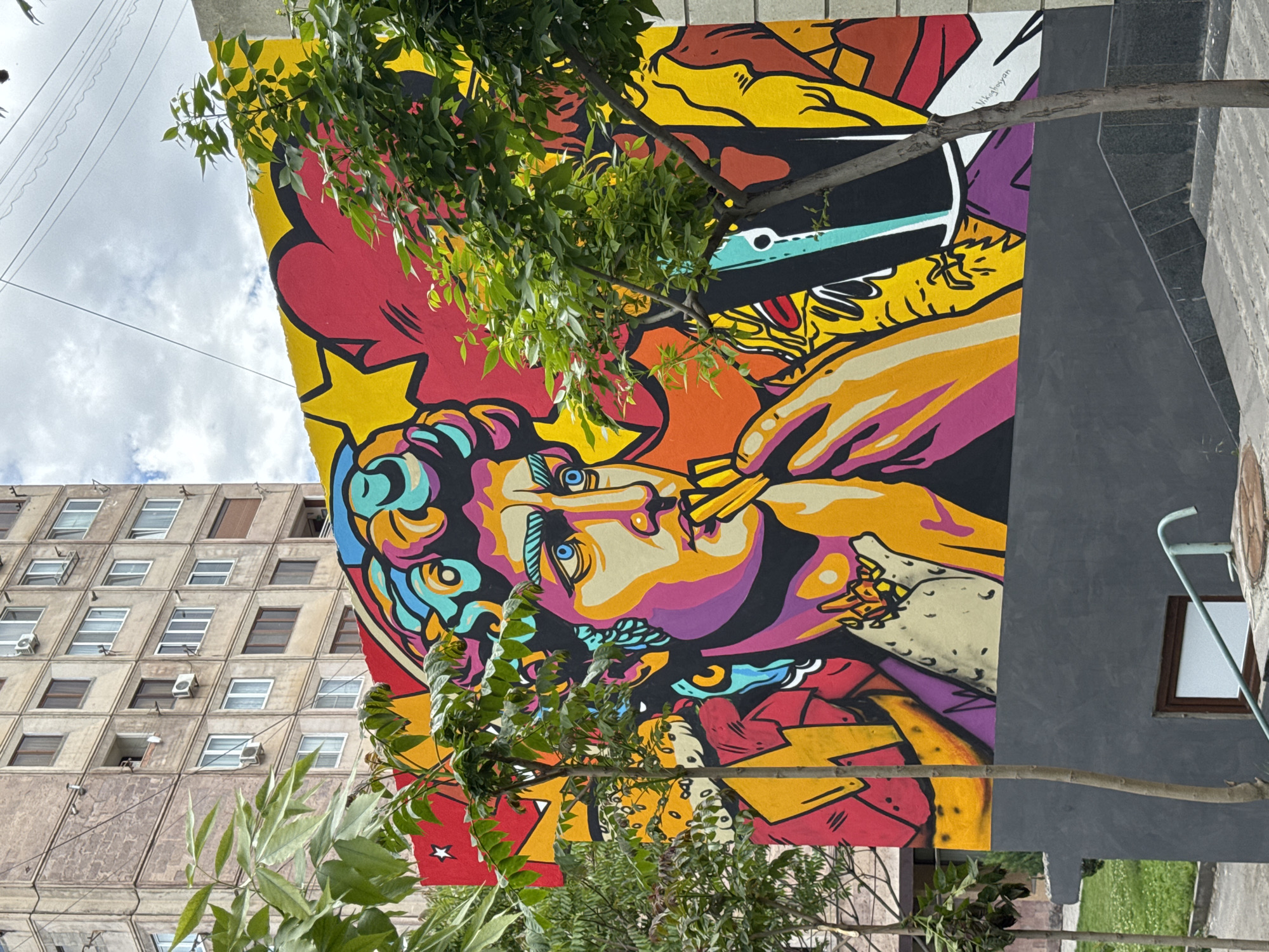 The image shows a vibrant mural in Yerevan, Armenia. The mural is painted on the side of a building and features a colorful, stylized portrait of a person smoking. The artwork includes bold lines, bright colors like yellow, red, orange, purple, and blue, and dynamic patterns. A tree with green foliage partially obscures the mural. A multistory building with a distinct façade of square windows is visible in the background. A gray wall is adjacent to the mural on the right.