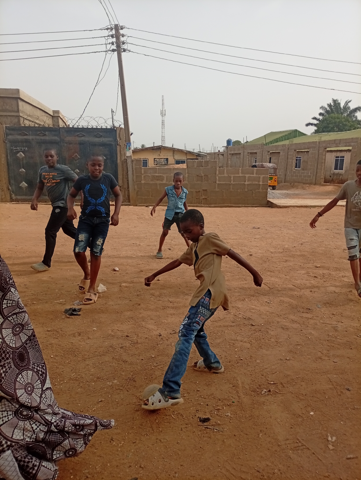 In Sabon Tasha, Nigeria, a group of five boys are playing on a dirt lot. The boy in the foreground wears a brown shirt and jeans, one foot raised as if kicking. A woman's patterned skirt is partially visible on the left. The background includes unfinished block buildings, a utility pole with wires, a gated compound, and a distant communication tower.