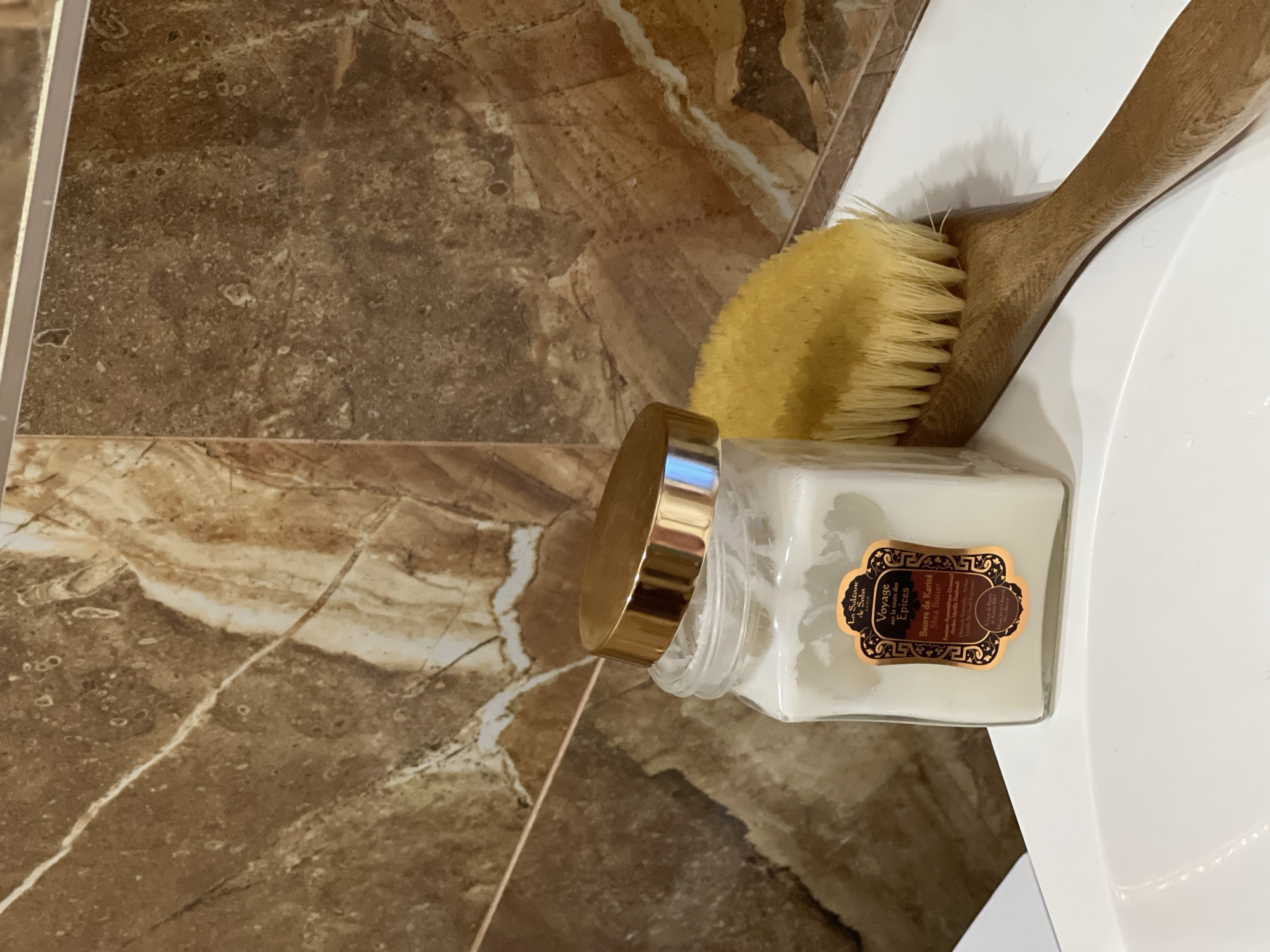 Here is a description of the image:
The image shows a bathroom scene. A partially visible white sink is positioned to the right, next to brown and tan marble-like tiles. On the sink's edge, there are two brushes: a bath brush and a smaller facial brush. The focus of the image is a clear glass jar with a golden lid. A label on the jar reads "La Sulzone de Saba Voyage sur la route des Epices Beurre de Karité Shea Butter Ayurvidlique". The jar sits near the brushes on the edge of the sink. The environment suggests a well-maintained bathroom.
I cannot provide specific location details for Khimki, Russia, as they are not depicted in the image.