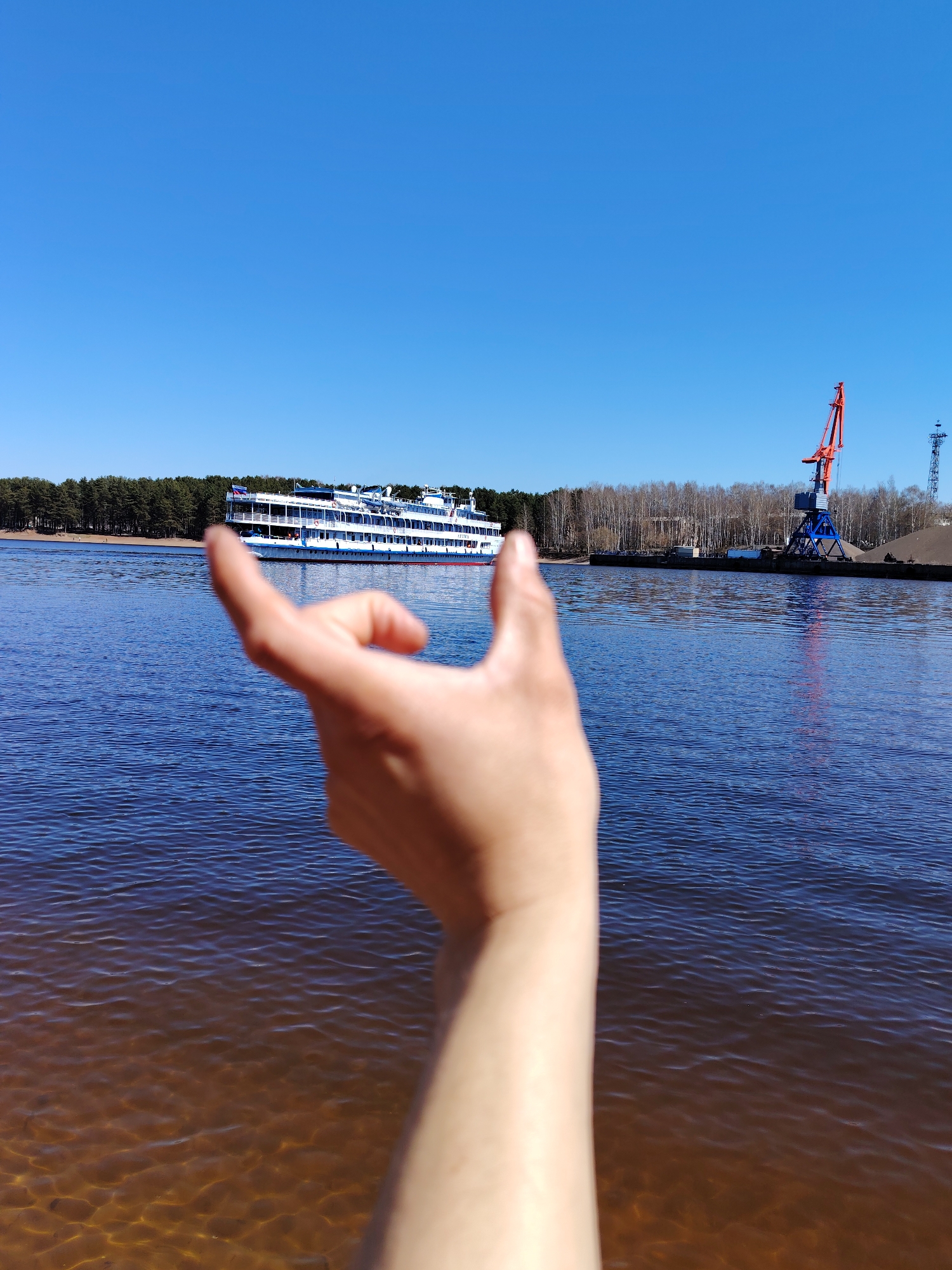 The image shows a river landscape with a large white passenger ship in the distance. In the foreground, a hand is partially obscuring the view. On the right side of the river, there is a harbor with a large red and blue crane and a pile of gravel. A line of trees is visible on the far bank of the river. The sky is clear and blue. Based on the image content, including the presence of a large river cruise ship and industrial harbor elements, this scene likely depicts a location along the Kama River in the city of Chaikovsky, Russia.