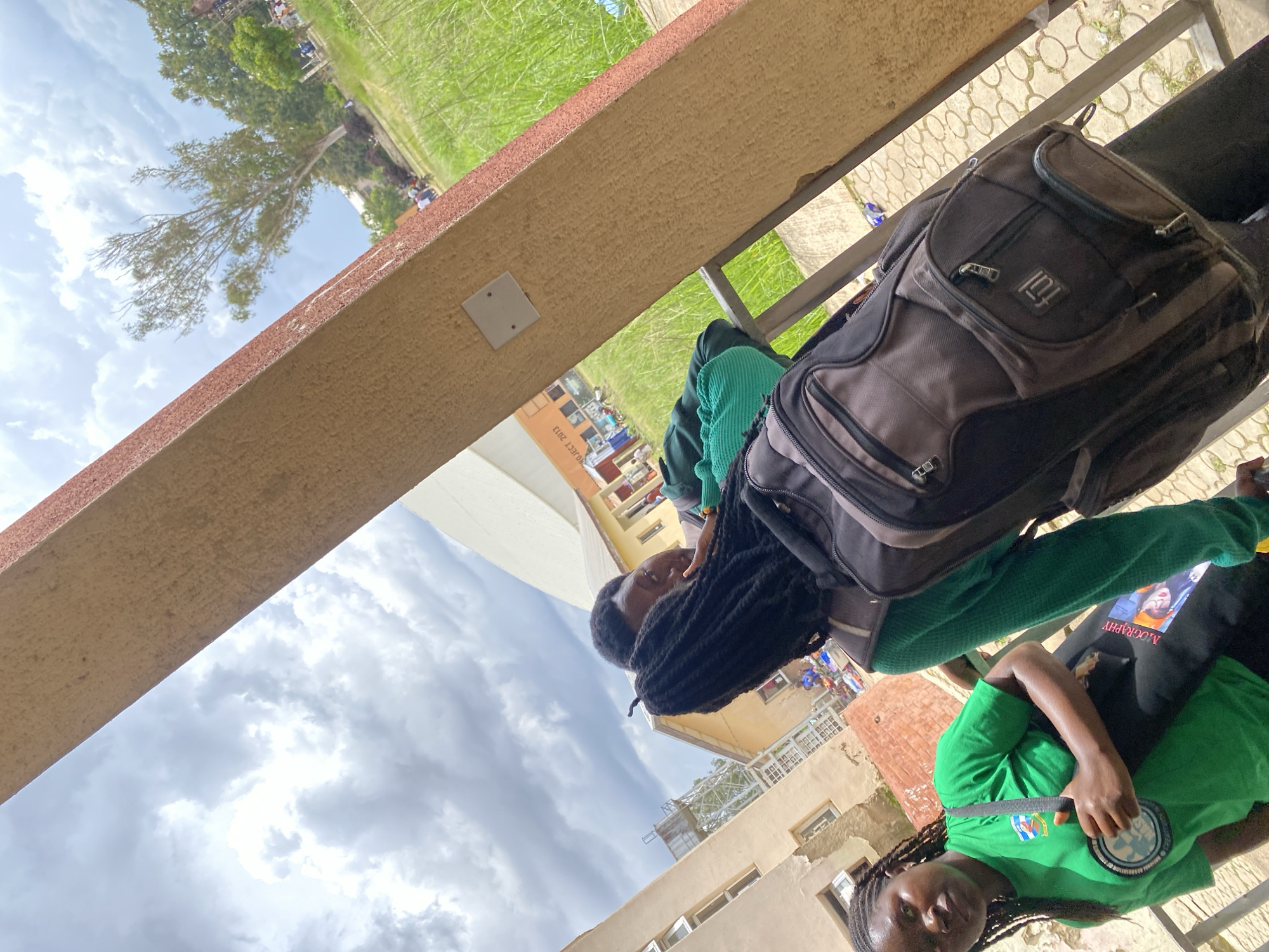 In Jos, Nigeria, two young Black individuals stand outdoors near a building. The person on the left, a woman, wears a bright green t-shirt with a circular logo on it and has braided hair. She is holding a black bag strap that goes across her body, with a black bag slung over her shoulder. To her right, another young Black person, with dark dreadlocks, is wearing a green sweater and a dark backpack. They are facing each other and appear to be interacting. The background reveals a building with windows and a water tower in the distance, along with a grassy area and trees. The sky is overcast with dramatic clouds.
