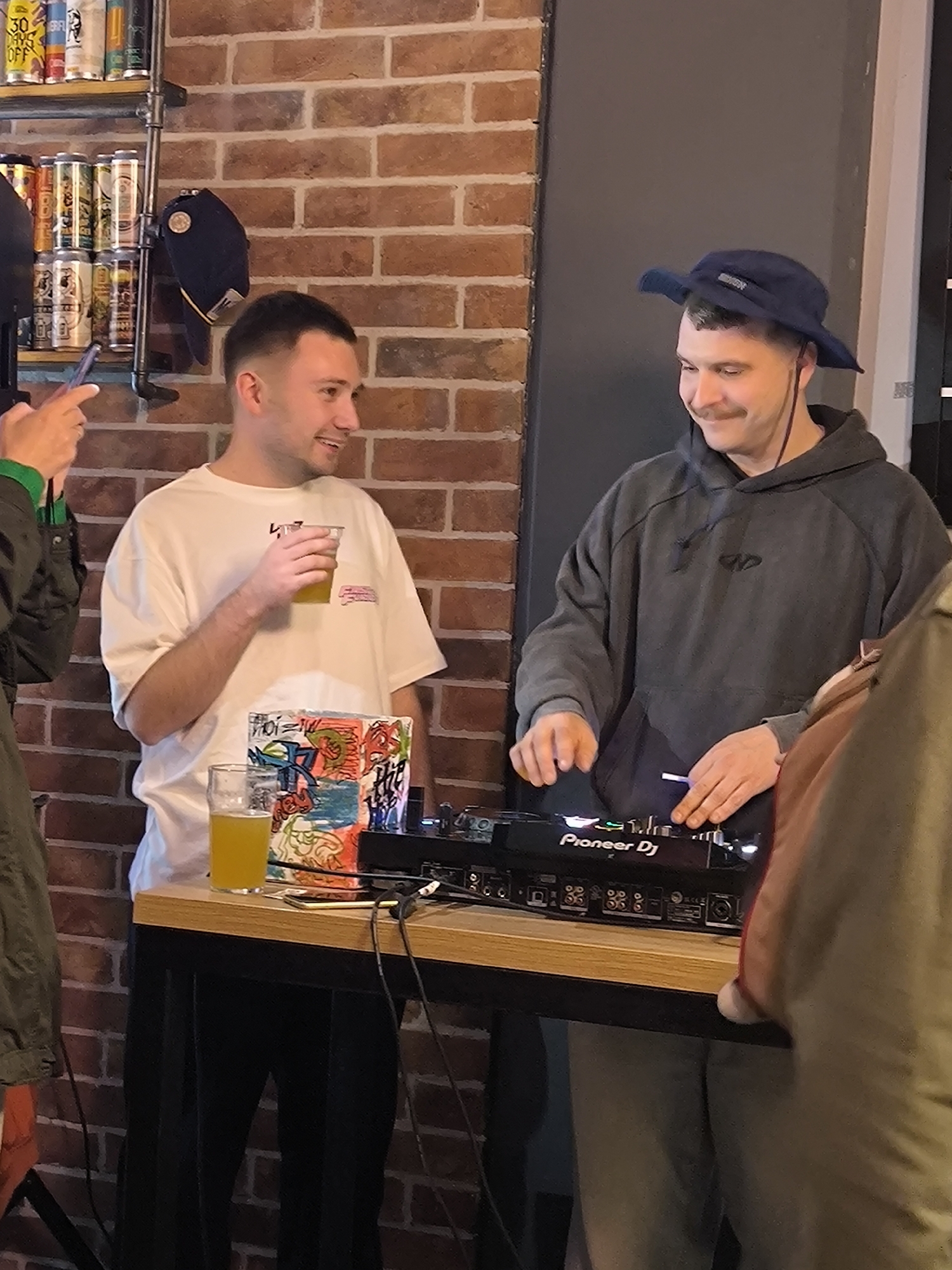 The image displays an indoor scene featuring three individuals within what appears to be a casual establishment. A red brick wall serves as the primary background on the left, while a dark grey panel or doorway is visible on the right.
In the center, a male subject with short dark hair and a white short-sleeved t-shirt stands smiling, holding a clear plastic cup with yellow liquid in his right hand. To his right, an adult male subject, presumed to be a DJ, wears a dark blue bucket hat and a dark grey hooded sweatshirt. He has a mustache and smiles faintly while operating a black "Pioneer DJ" branded mixer or controller with both hands. This equipment rests on a light wooden tabletop. A tall, clear glass containing yellow liquid is also on the table, positioned to the left of the DJ equipment, beside a multi-colored, graffiti-style art object.
On the far left, partially visible, a third individual holds a smartphone, pointed towards the DJ and central subject, possibly recording or photographing. A metal shelving unit, constructed from pipes, is affixed to the brick wall above the central subject. This unit holds multiple rows of various aluminum beverage cans, and a dark blue baseball cap hangs from a lower pipe. The rightmost edge of the image shows a partial view of a fourth individual's shoulder and arm, clad in a light brown garment, partially obscuring the DJ's lower body. The exact location within the city of Bar, Montenegro, is not identifiable from the visual information provided.