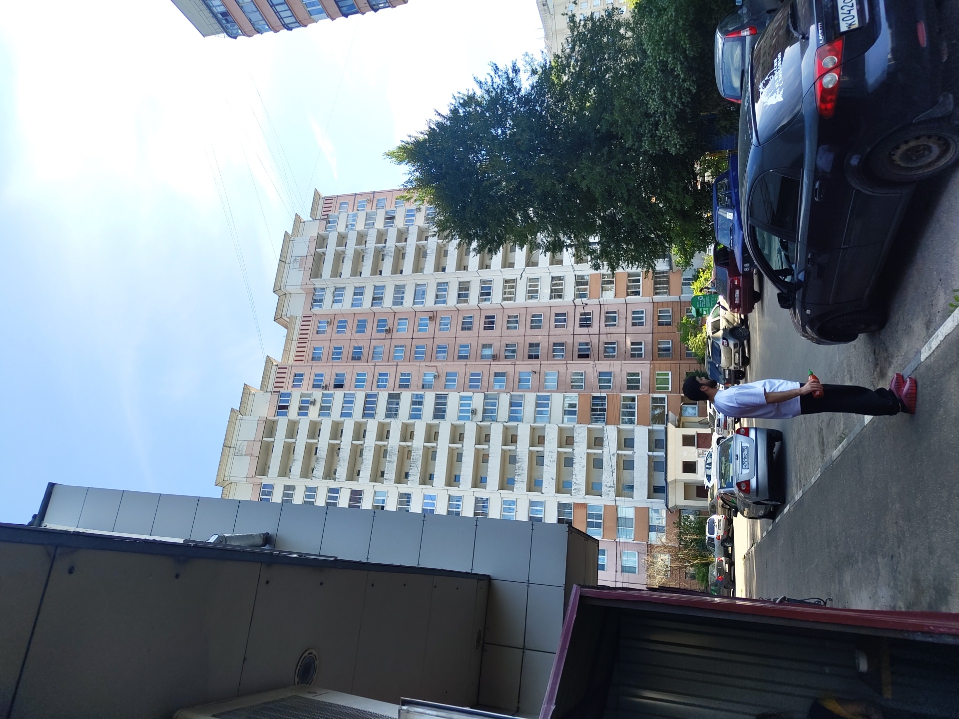 A man in a white t-shirt and black pants stands on a paved surface, holding a red object in his right hand. He is facing to the left. Behind him, several parked cars are visible. In the background, a large, tall apartment building with numerous balconies and windows dominates the scene. A dense green tree is located to the right of the building. The sky is clear and blue, with some faint cloud streaks. The setting appears to be an urban environment, likely in the city of Zhukovsky, Russia, given the context provided.