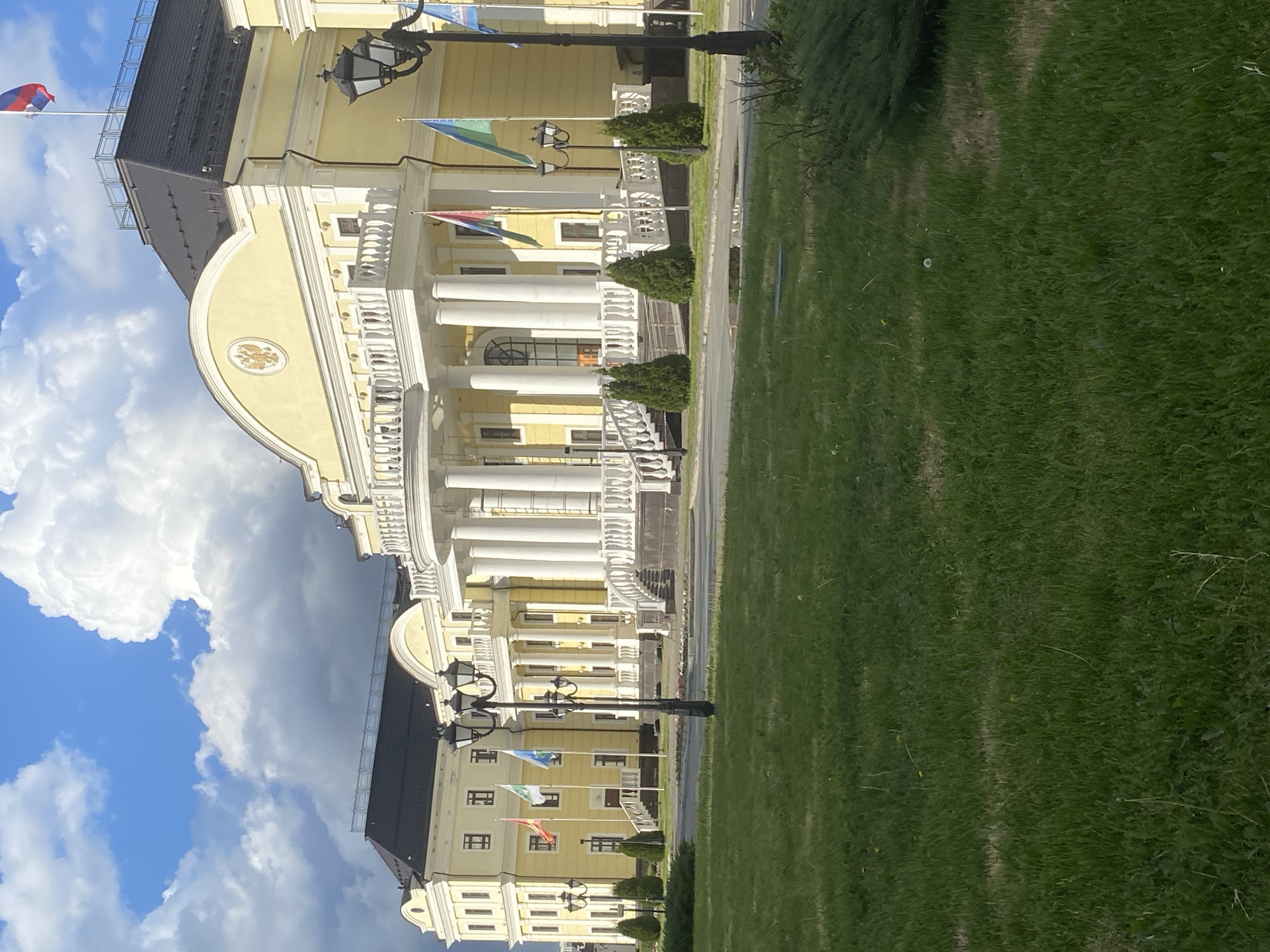 The image depicts a yellow building with white columns, likely a government or public building, situated in Yekaterinburg, Russia. The building features a black roof and flies several flags from its façade. A paved area and a green lawn surround the building. The sky is blue with white clouds. Streetlights stand along the edge of the paved area.