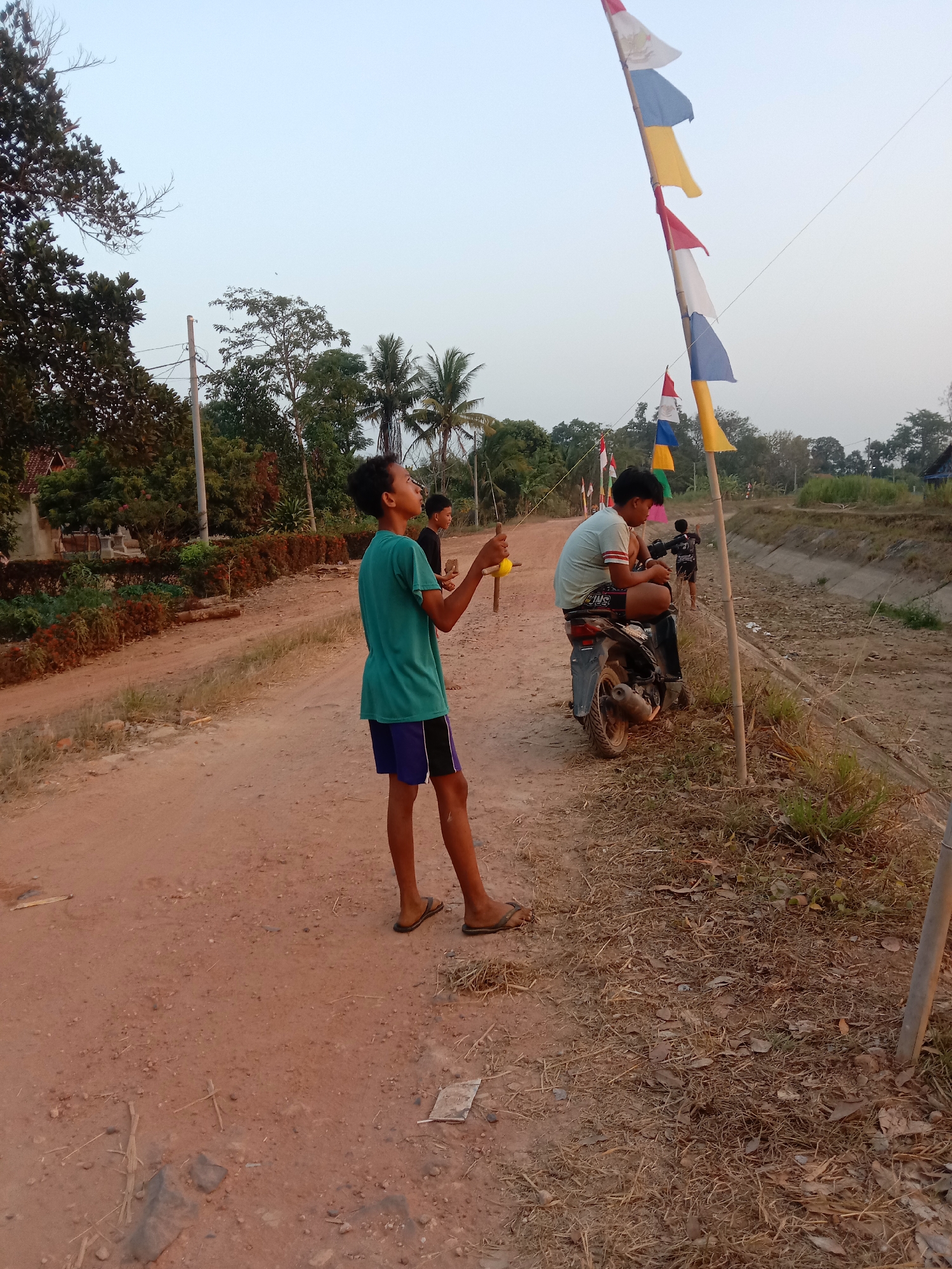 A boy in a teal t-shirt and blue shorts stands on a dirt road, holding a kite string. Behind him, another boy sits on a motorcycle. In the background, a line of flags on bamboo poles flutters in the breeze. The scene appears to be in a rural area, possibly on the outskirts of a city. The sky is overcast, and the overall lighting suggests it is late afternoon or early evening. The dirt road is lined with vegetation, including trees and some dry grass. A drainage ditch runs alongside the road. There is a house visible in the far left background.