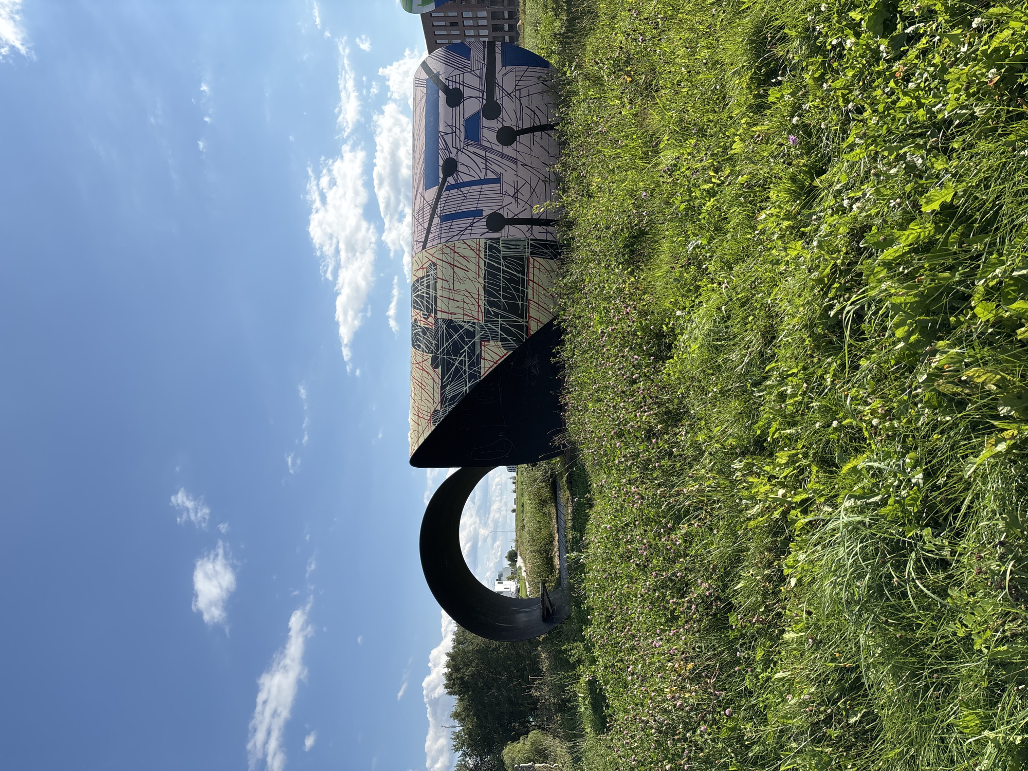 A large, abstract metal sculpture is situated in a grassy field under a blue sky with scattered white clouds. The sculpture is cylindrical and appears to be a segmented or broken form. One section is a large, dark, curved arch. The other section is a more complex, elongated form covered in a colorful, graphic pattern of lines, grids, and dark circular shapes. The grass in the foreground is tall and green, with some small pink and white wildflowers. In the background, there are trees and modern buildings. The scene is well-lit by natural sunlight.