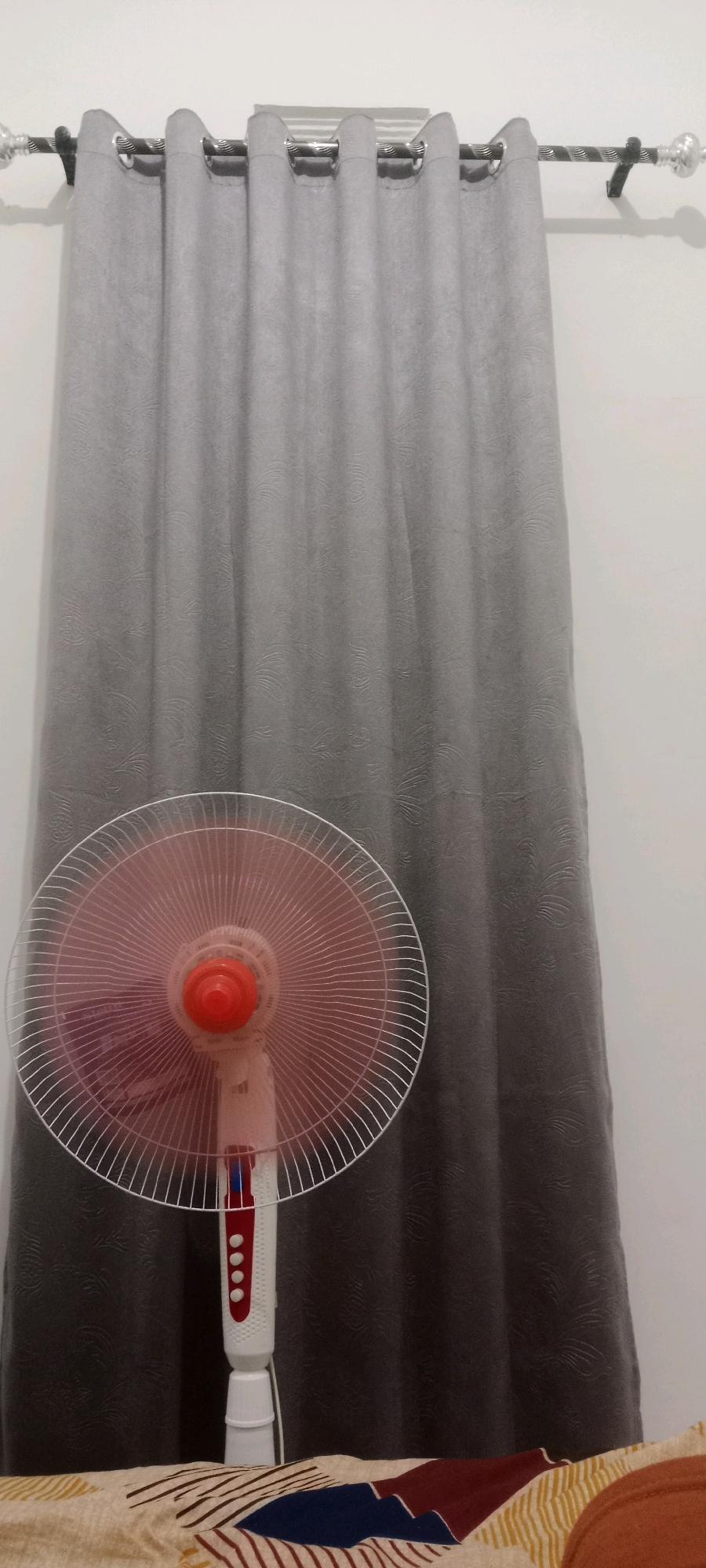 A floor fan with a pink motor housing and a white base is positioned in front of a grey curtain. The fan's grille is visible, and the curtain appears to have a subtle textured pattern. The scene is indoors, with a plain wall visible behind the curtain. The lower portion of the image shows a patterned surface, possibly a bedspread. There are no people present in the image. The location is identified as Serongga, Indonesia.