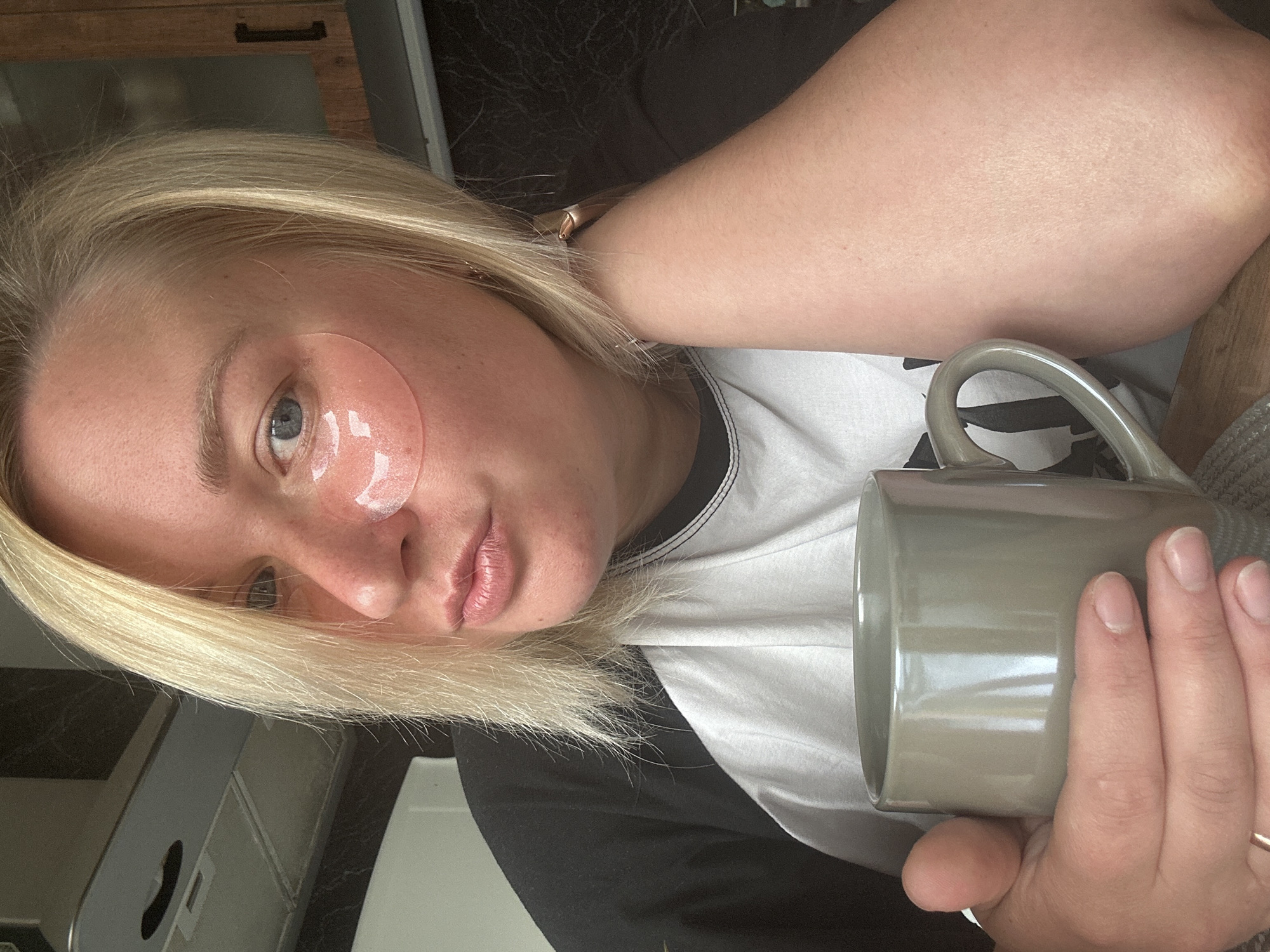 A close-up, waist-up, frontal view shows a fair-skinned, blonde-haired woman with blue eyes and pink under-eye patches, holding a grey mug of coffee. She is wearing a black and white t-shirt. Her right arm is bent, with her hand resting on her neck and her forearm visible. The background is blurred, showing kitchen cabinetry and what appears to be a stove hood. The scene suggests an indoor domestic setting, likely a kitchen or dining area. There is no information in the image to indicate the location is Chaikovsky, Russia.