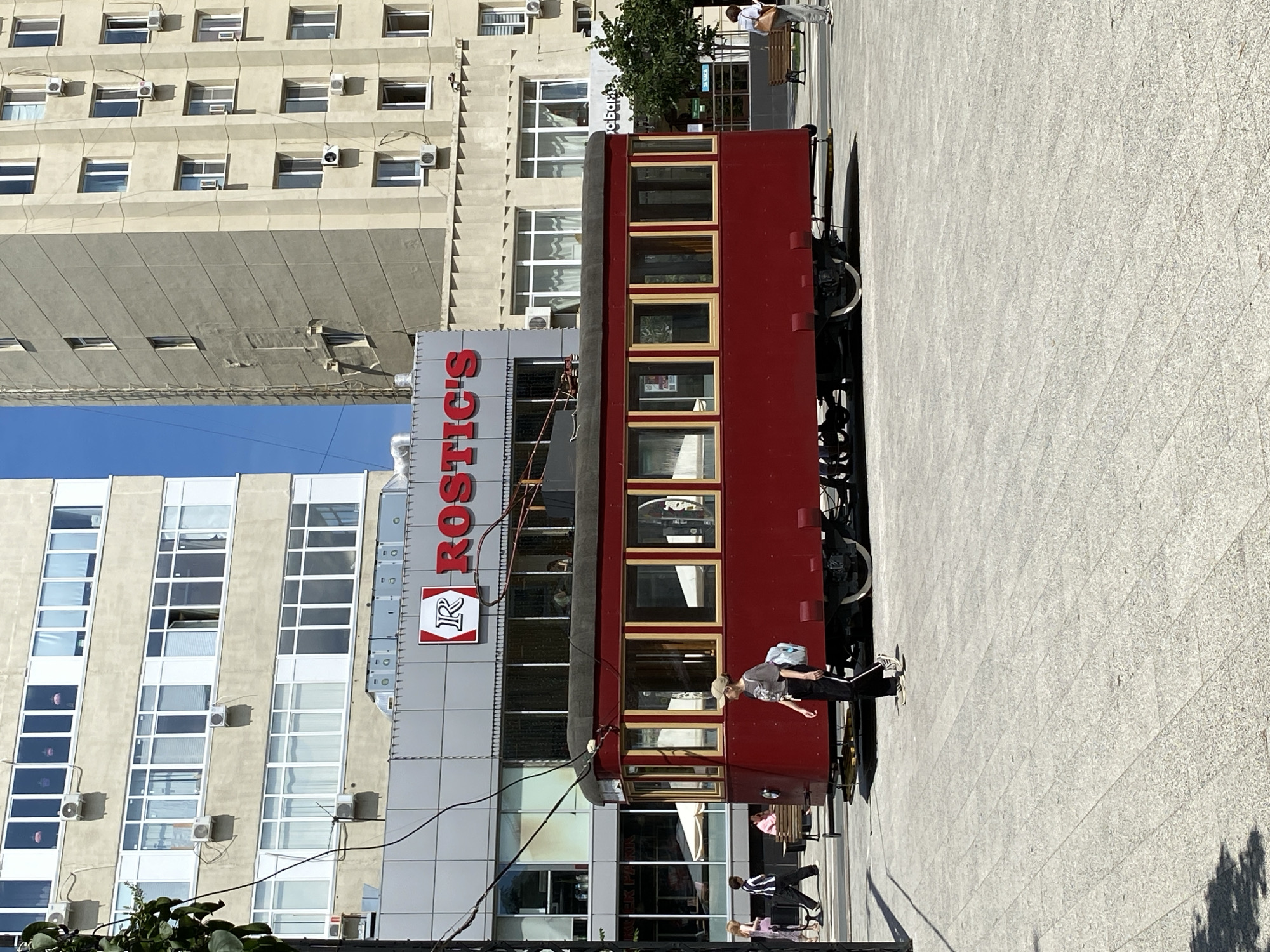 The image shows a red vintage tram parked on a paved plaza in front of a multi-story building. The building has a modern facade with numerous windows and a prominent sign that reads "ROSTIC'S" in large red letters, with a logo below it. The tram is old-fashioned with numerous windows and wooden trim.
Several people are visible in the scene. One person, wearing a cap and a backpack, is walking near the tram. Further in the background, a few other individuals are seen, some walking and one sitting on a bench. The setting appears to be a sunny day, indicated by the clear blue sky and the shadows cast on the ground. The tram's presence suggests it might be a stationary attraction, a restaurant, or part of a historical display. The overall atmosphere seems relaxed and casual, typical of an urban public space. The building suggests a commercial or residential area.