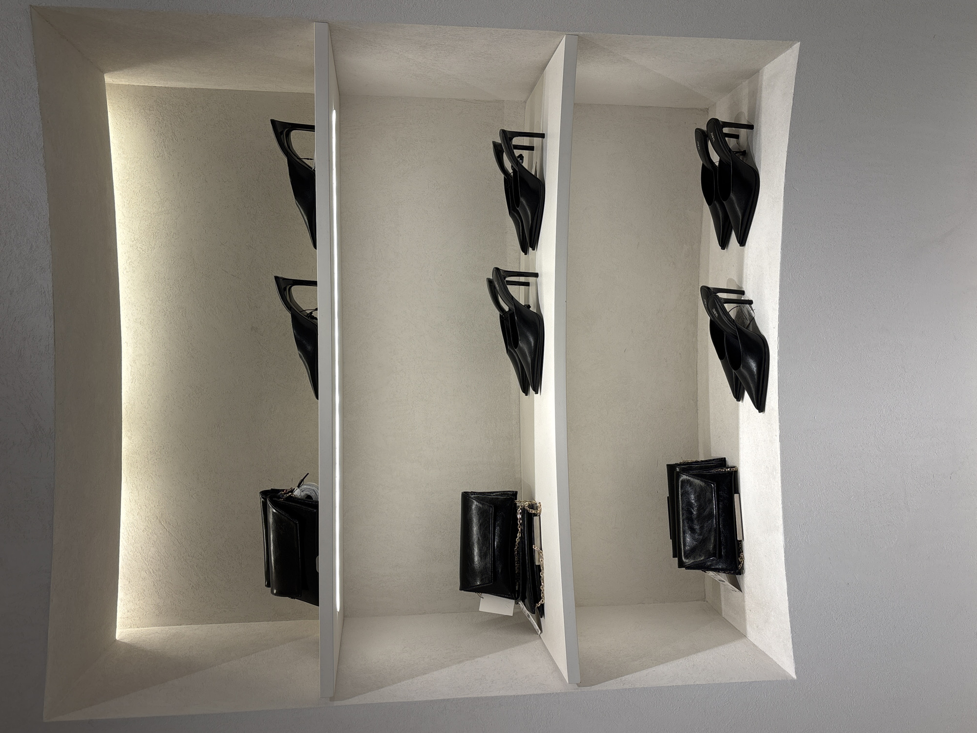 The image depicts a recessed display case with three compartments, likely within a retail store. The case is predominantly white, with textured walls and dividers. Each compartment contains black leather items. The upper shelves hold pairs of black high-heeled mules mounted to the wall by their heels, while the lowest shelves contain small black handbags. The image focuses on the display and lacks any identifiable information about the specific location in Sofia, Bulgaria. No people or interactions are visible. The environment consists solely of the display case and the surrounding wall.