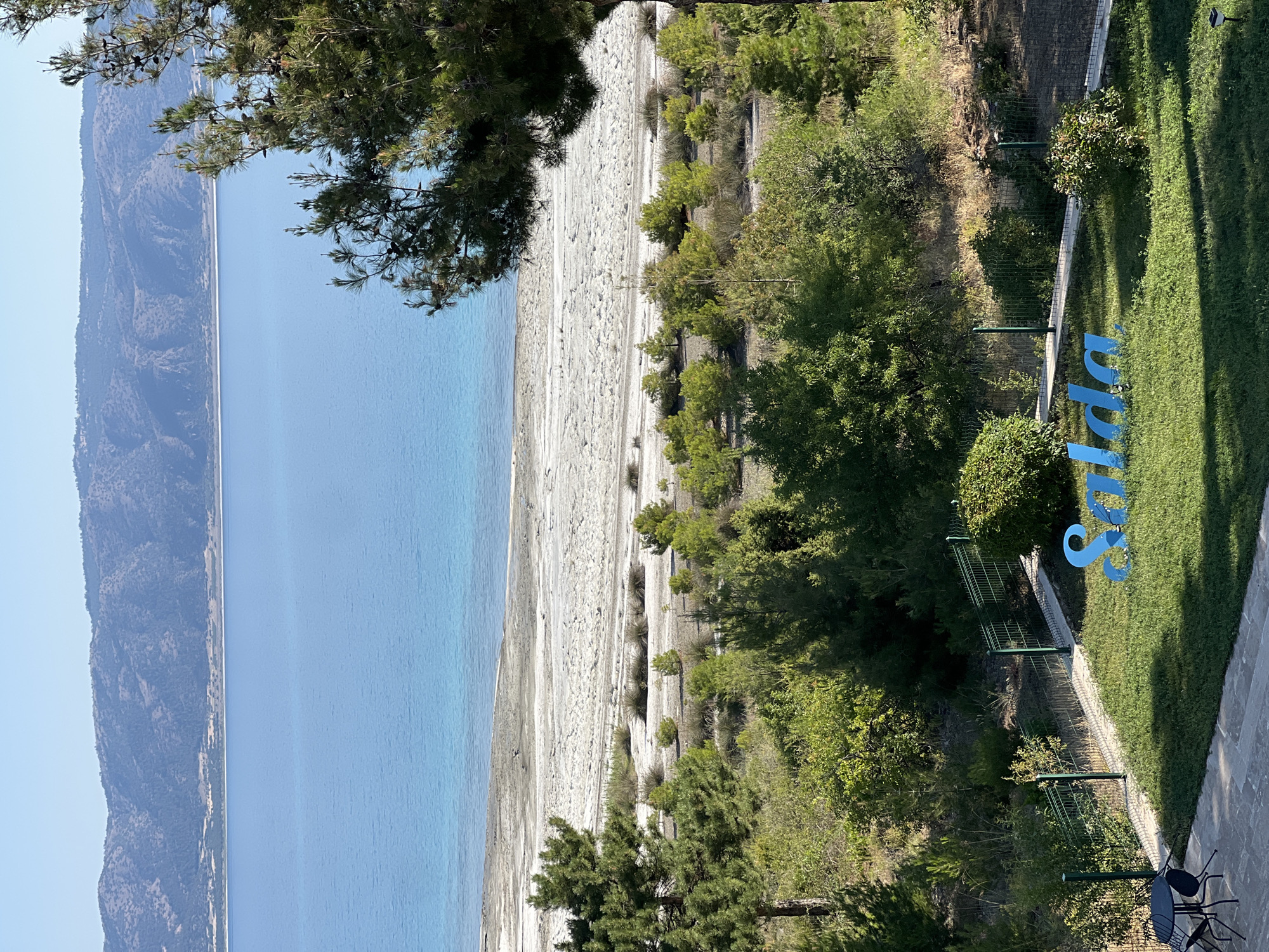 The image shows a scenic view of Salda Lake in Yeşilova, Turkey. In the foreground, a manicured green lawn features a large, blue, three-dimensional sign spelling out "Salda". A black metal table and chairs are set up on a paved patio to the left. Behind the lawn, a green metal fence borders an area with lush green trees and bushes, leading up a gentle slope.
The middle ground consists of a sandy, somewhat barren area with sparse vegetation, bordering the lake. The lake itself dominates the background, displaying a vibrant blue hue that transitions to a lighter turquoise near the shore. In the distance, rolling hills with sparse vegetation and trees form the horizon under a clear blue sky. The overall scene suggests a well-maintained tourist area with a focus on the natural beauty of Salda Lake.