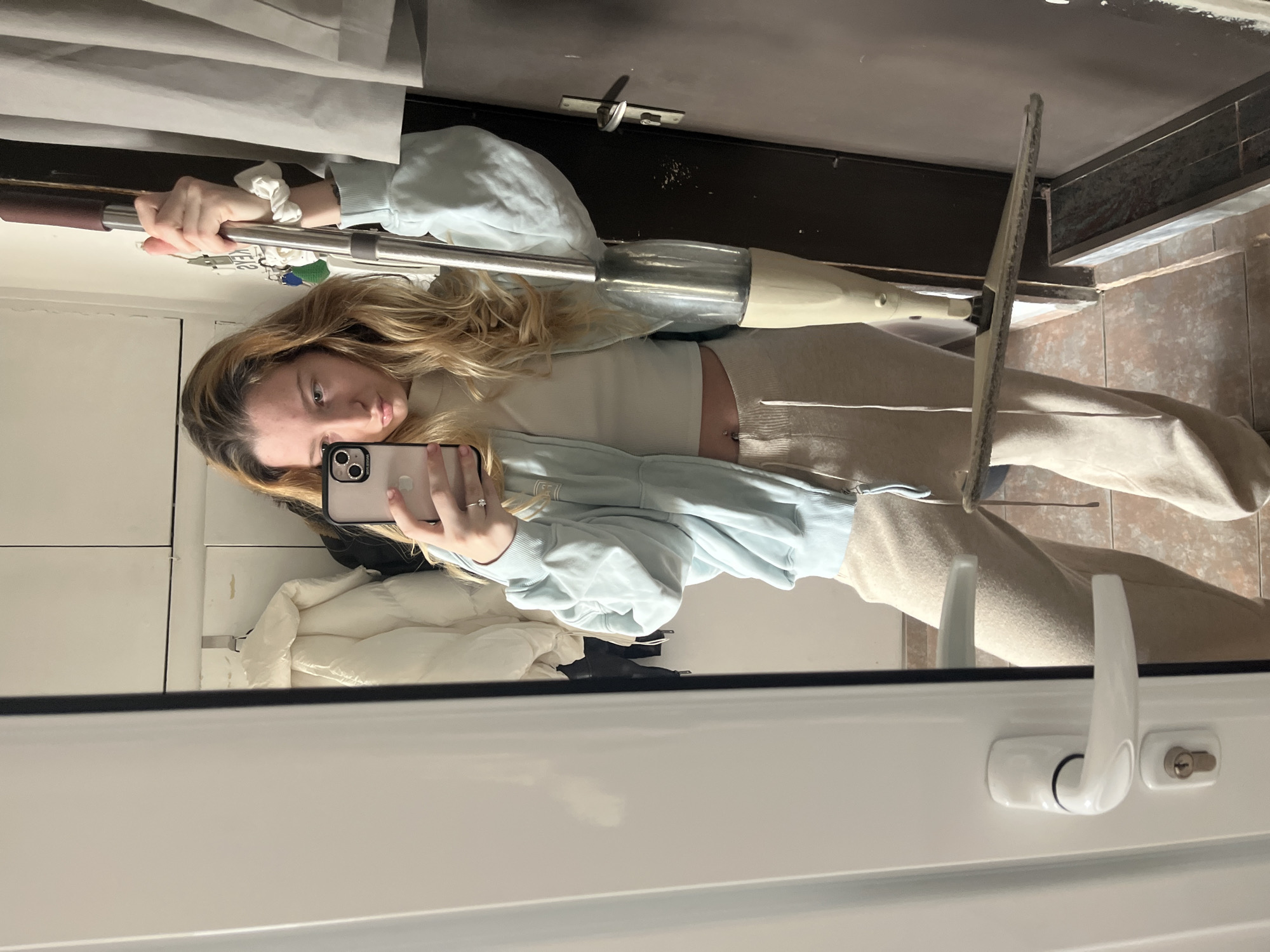 A mirror selfie depicts a young adult female in an indoor setting, identified as Varna, Bulgaria. The subject, possessing long blonde hair, is reflected from the mid-thigh up. She holds a silver smartphone in her left hand, positioned to capture the image. Her right hand grasps the upper pole of a beige and silver floor spray mop, holding it upright with the cleaning head near the floor. She is attired in a light blue zipped hoodie over a beige high-neck cropped top, revealing a navel piercing, and beige sweatpants. A ring is visible on her right ring finger, and a white scrunchie is on her right wrist. In the reflected background, to the subject's left, a white padded jacket and a dark garment hang on hooks against a light wall or cabinet. To her right, a dark brown door or wall features a metal key holder with several keys attached. The floor is covered with light brown square tiles. A white door frame, partially open door, and a white handle with a keyhole are visible on the left side of the reflection. A narrow metal grate or vent is present on the floor against the dark wall on the right.