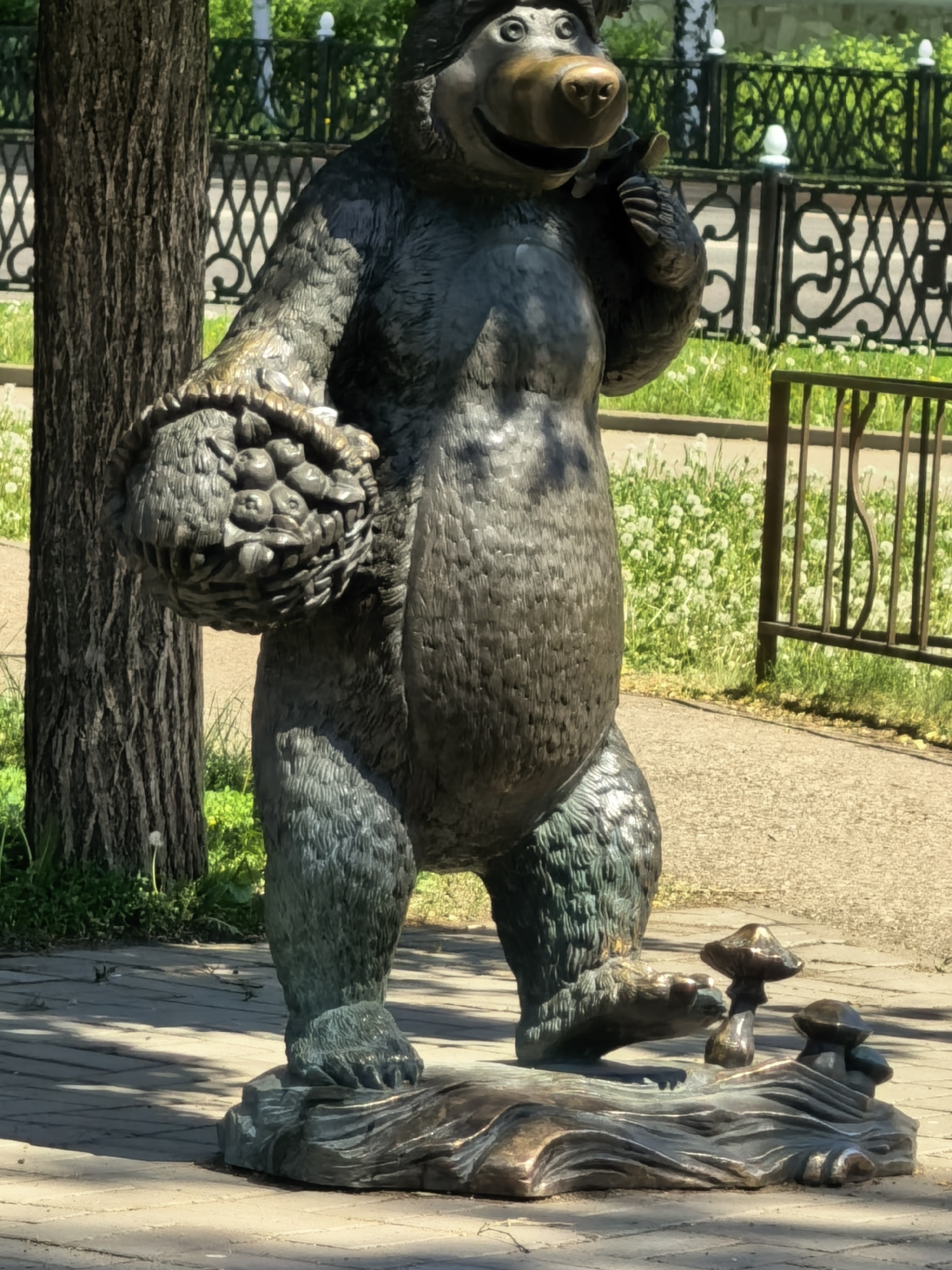 A bronze statue of a bear stands on a paved walkway, with a basket of fruit in its left arm. The bear stands on a base adorned with mushrooms. A tree trunk is on the left side of the statue, with a dark metal fence in the background. The photograph was taken in Sterlitamak, Russia.