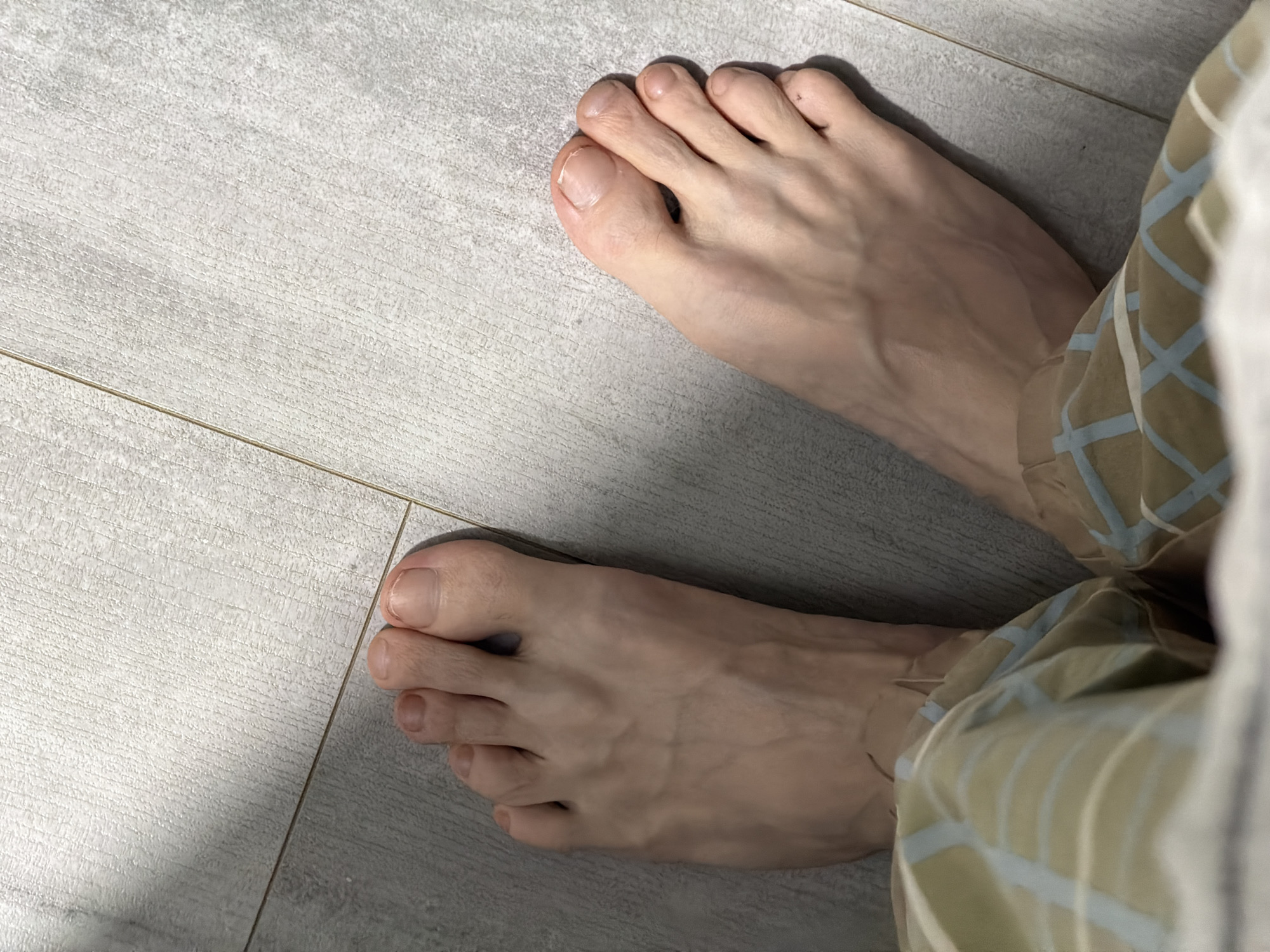 The image shows a pair of bare feet on a light gray tiled floor. The feet are positioned with the toes pointing towards the top of the frame, and the soles facing upwards. Sunlight casts shadows across the tiles. The feet are fair-skinned and appear to be those of an adult. They are partially obscured by a pair of light-colored pants with a grid pattern of light blue and beige lines. The scene appears to be indoors, in a domestic setting, possibly a bathroom or bedroom. There is no indication of the location being in the city of Naberezhnye Chelny, Russia.
