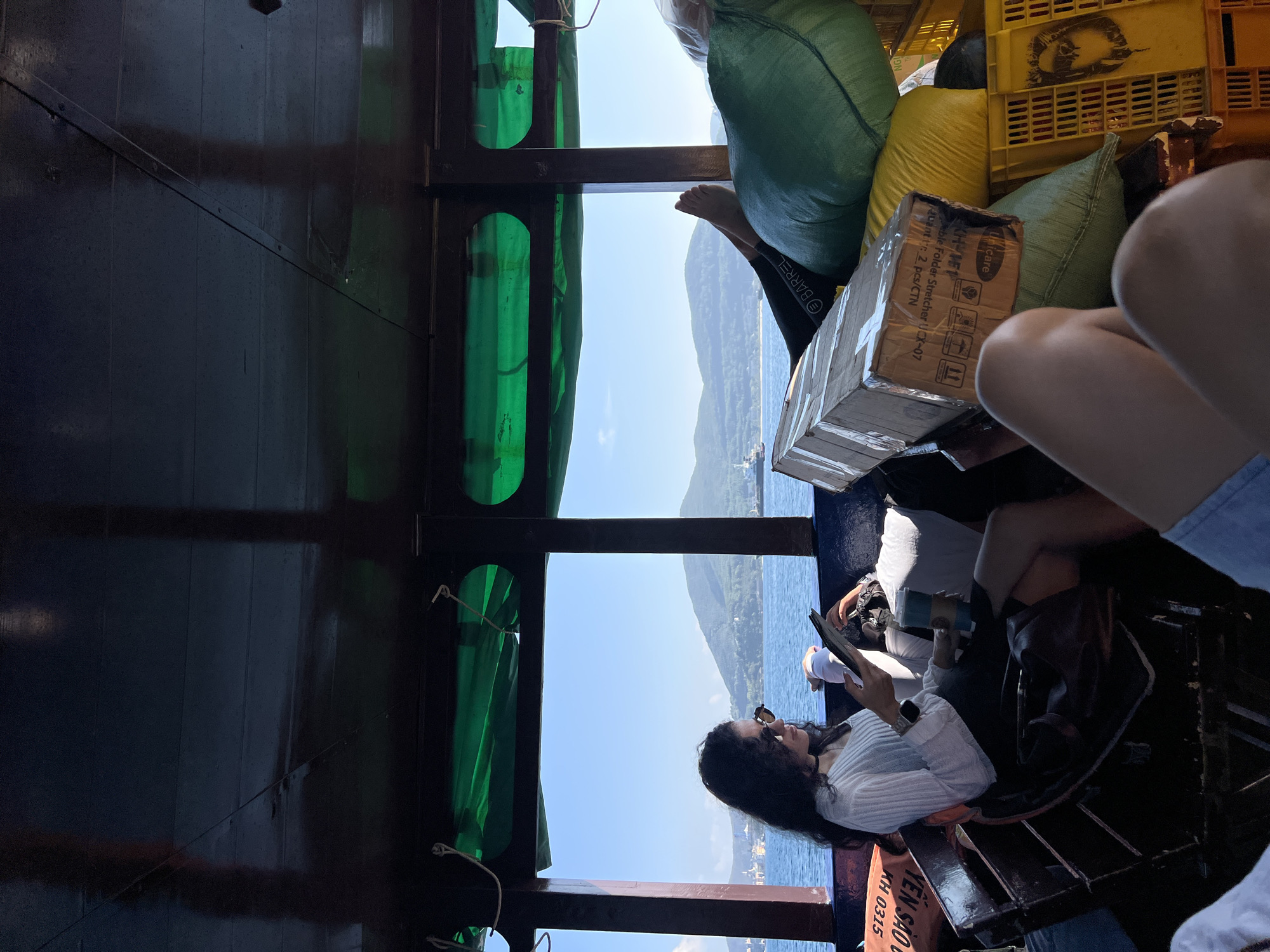 The image appears to be taken inside a boat, possibly in Bac Nha Trang Ward, Vietnam, based on the text "YÊN SÀO KH 0315" and the coastal scenery visible through the window. A woman with dark hair and sunglasses is seated, looking at a tablet or phone. There are bags and yellow crates stacked nearby, suggesting transport of goods. The water and mountains are visible in the background through the boat's window, indicating a coastal or island location. The boat's interior is dark wood. There is also a cardboard box labeled "Uni nicare" with text indicating "Foldable Folder Stretcher UCX-07" and "Quantity: 2 pcs/CTN".