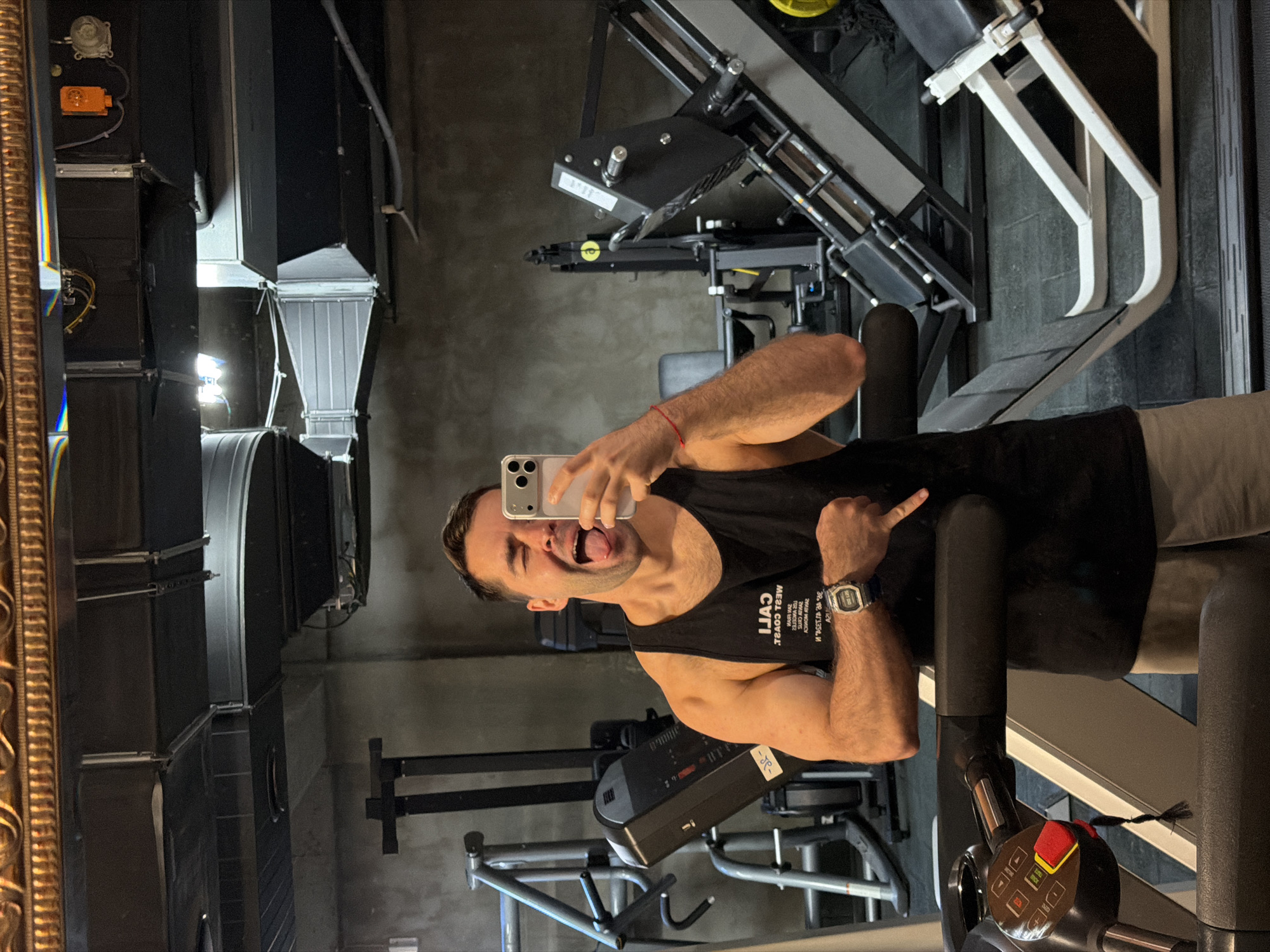 The image is a selfie taken in a gym, likely in Tbilisi, Georgia. A man is holding a smartphone and sticking out his tongue. He is wearing a black tank top and a wristwatch. The gym has various exercise machines, including a rowing machine, a leg press, and ventilation ducts. The background features neutral grey walls, and a corner of a decorative mirror is visible on the left.