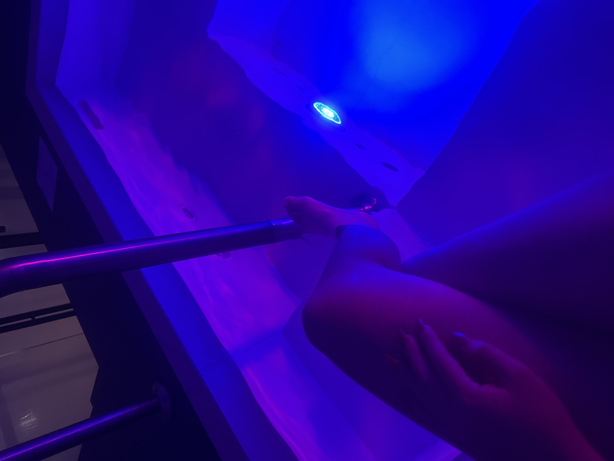 The image shows a person in a sensory deprivation tank, likely a floatation therapy session. The tank is illuminated with blue and purple light, creating an otherworldly atmosphere. The person is lying down in the water, with their hand resting on their leg. A metal bar runs along the side of the tank, likely for support. The exact location within Huixquilucan de Degollado, Mexico is not discernable from the image.