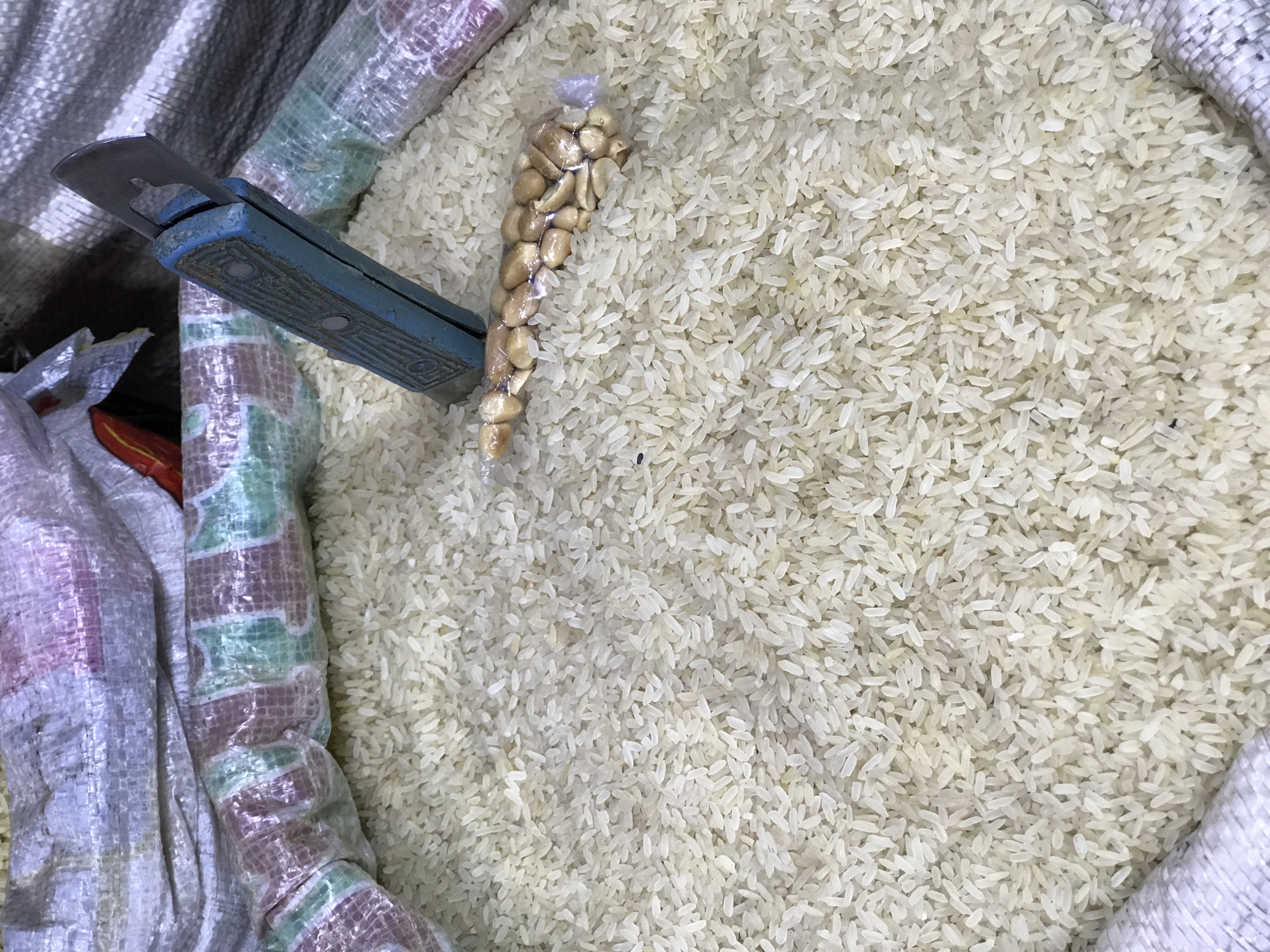 This close-up, top-down shot, taken in Abuja, Nigeria, captures a large quantity of loose white long-grain rice, likely contained within a sizable woven polypropylene sack. The rice grains fill most of the frame, appearing consistent in size and color.
On the left side of the image, a blue-handled utility knife or sack opener is partially inserted into the rice, with its worn metal blade and textured, faded blue handle clearly visible. Adjacent to the knife, a small, clear plastic bag filled with shelled peanuts rests on the surface of the rice.
Portions of the patterned, possibly woven, material of the main rice sack and other similar storage containers are visible along the left and top edges of the frame, suggesting a market stall or a bulk storage area. The overall scene indicates the selling or distribution of staple food items in a local market environment. No people or visible text are present in the image.