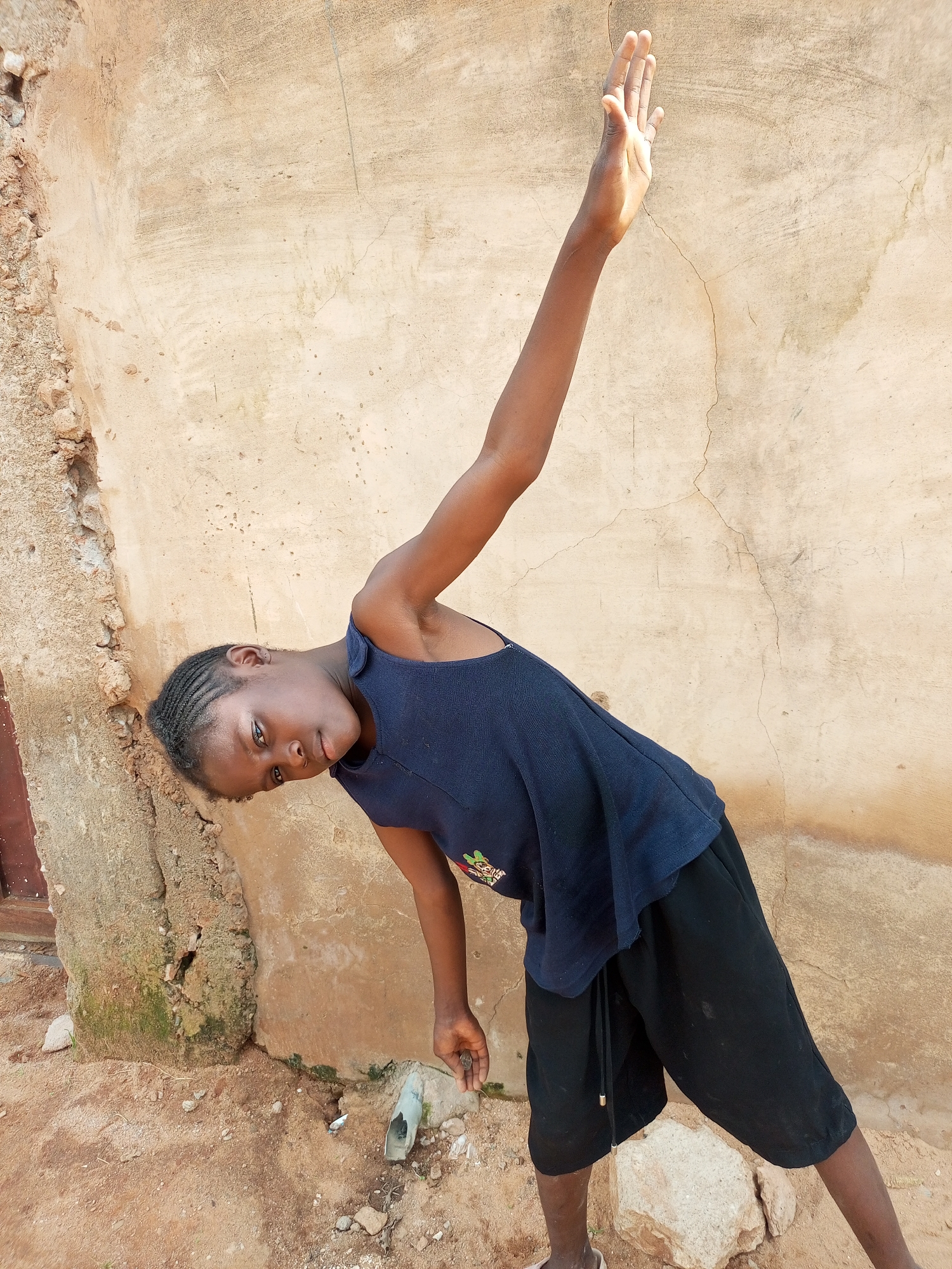 A young African child with braided hair is leaning to their left while posing for a photo in front of a textured, beige wall. The child wears a dark blue sleeveless top with a design on the front and black shorts. Their right arm is extended upwards and slightly back, with their palm open. The ground is a mix of dirt and rocks, with some vegetation and a small drainpipe visible. A portion of a building or wall with rough, crumbling texture is on the left side of the image. The location is identified as Tempure, Nigeria.