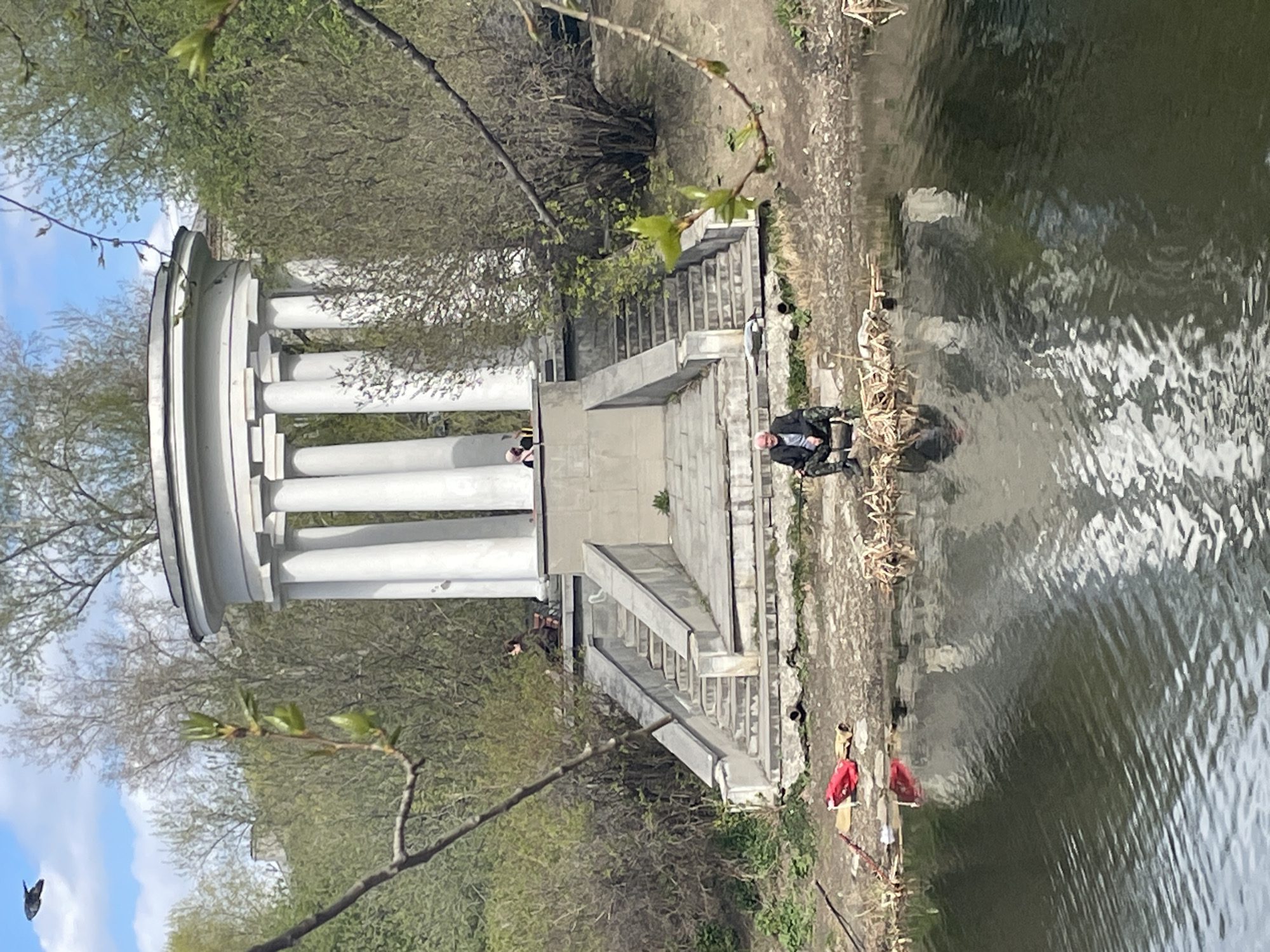 The image shows a view of the Rotonda, a white columned gazebo, located in the Kharitonov Garden park in Yekaterinburg, Russia. The Rotonda sits at the top of stone steps leading down to a body of water, possibly a pond or a section of the Iset River that runs through the park.
A man is sitting on the bank near the water. Other people are on the steps leading up to the Rotonda. The environment includes greenery, trees, and the water surface. The sky is visible in the upper left.