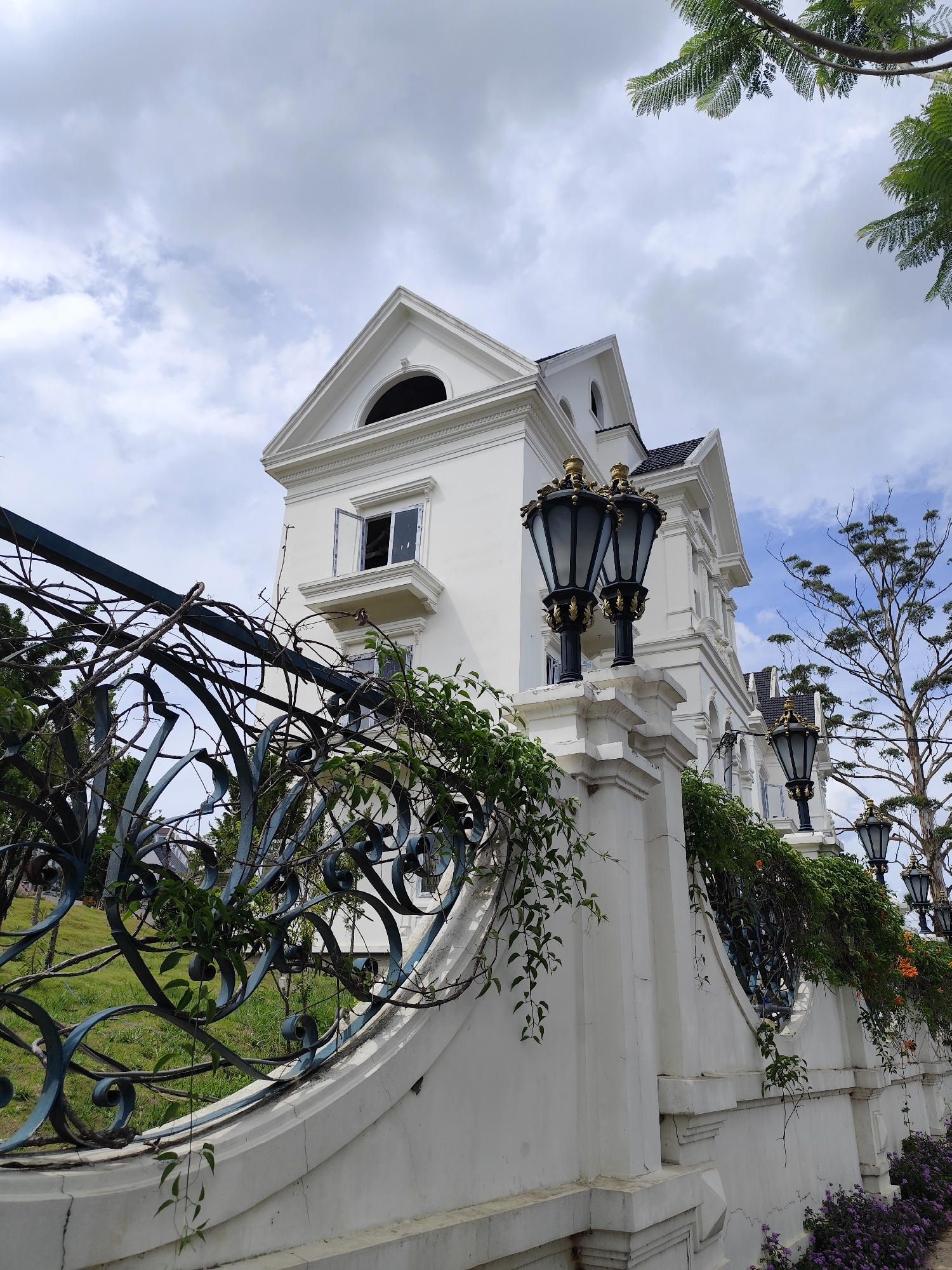 Captured in Lâm Viên, Đà Lạt, Vietnam, a white, multi-story building stands behind a curved, wrought-iron fence adorned with climbing plants. Black ornate street lamps are mounted on the fence's supporting posts. A partially open window is visible on the building's upper level. The sky is cloudy. Beyond the fence, greenery is apparent.