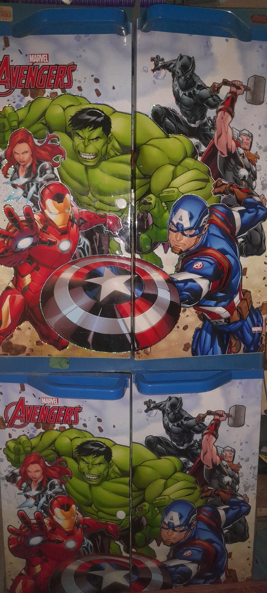 The image shows a close-up of a printed graphic on what appears to be a storage container, likely a toy box or a drawer unit, featuring the Marvel Avengers characters. The graphic is split vertically down the middle. The left side displays Iron Man in the foreground, with Captain America's shield partially visible in front of him. Behind Iron Man are Black Widow and the Hulk. The right side features Captain America in the foreground, wielding his shield. Behind him are Black Panther, and Thor holding his hammer. Both halves of the graphic are identical in content and arrangement. The printing has a glittery effect, particularly noticeable on the characters' outlines and the "Avengers" logo at the top left. The upper portion of the image shows a blue plastic edge and handle, suggesting the container has a lid or is part of a larger furniture piece. The lower portion of the image shows another identical graphic, suggesting the container has multiple levels or drawers. The scene is indoors, with no discernible environmental details of the city of Tegal, Indonesia.