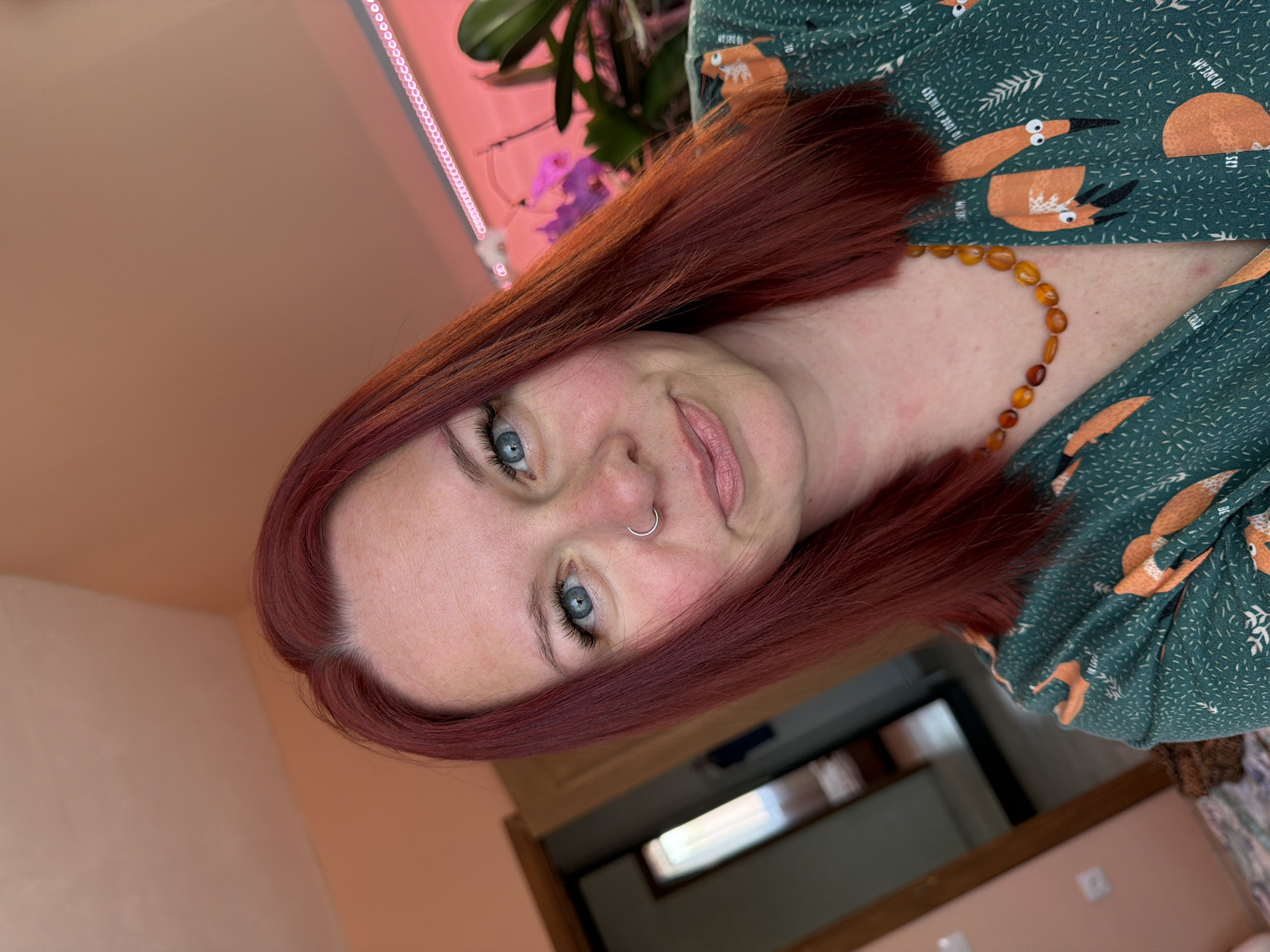 A close-up, slightly rotated image depicts a fair-skinned woman with red hair, blue eyes, and a silver nose ring. She is wearing a green top with a pattern of orange bird-like figures and a necklace of orange beads. The background includes a light pink wall, part of a window frame, and a plant with pink flowers. The location is unknown and cannot be verified from this image.