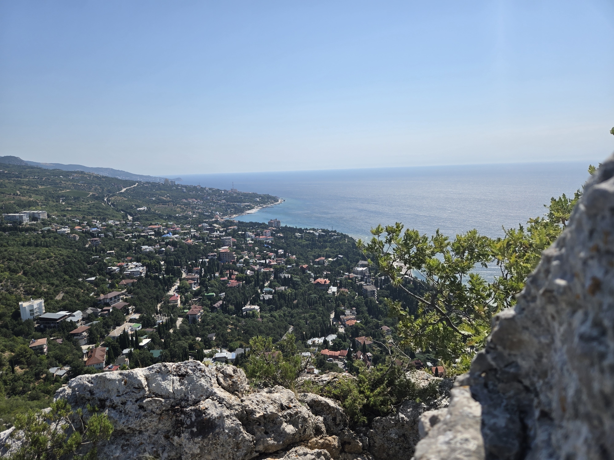The image captures a scenic overview of a coastal town nestled between lush green hills and the vast expanse of the sea. The foreground is dominated by rugged, light-colored rocks and a few green branches, suggesting a vantage point from a cliff or elevated terrain.
In the mid-ground, a densely populated town unfolds with numerous buildings, many featuring red-tiled roofs, interspersed with abundant trees. Winding roads are visible, connecting different parts of the settlement. The architecture appears to be a mix of residential houses and larger buildings, possibly hotels or apartment complexes, particularly closer to the coast.
The background showcases the clear blue sea meeting the horizon under a bright, cloudless sky. A distinct shoreline curves along the edge of the town, with what appears to be a beach area. The overall impression is of a sunny, tranquil day in a picturesque coastal area. The location is identified as Симеизский поселковый совет, Ukraine.