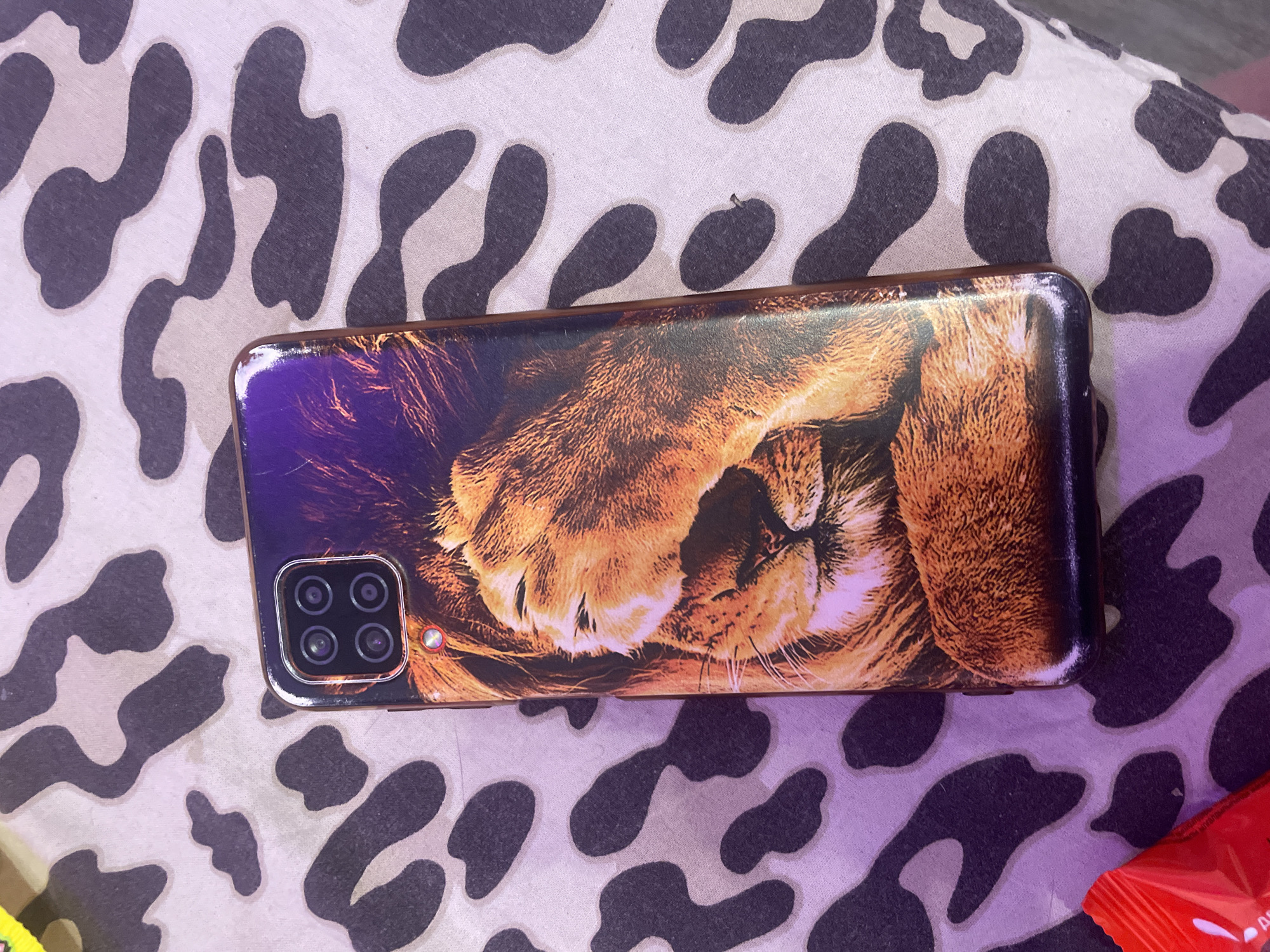 A phone case with a lion's face is placed on a fabric with a leopard print. The phone case is oriented vertically and the lion's face is in focus. The leopard print fabric is mostly out of focus. There is also a red object in the lower left corner of the image. The image was taken in the city of Irinovka, Russia.