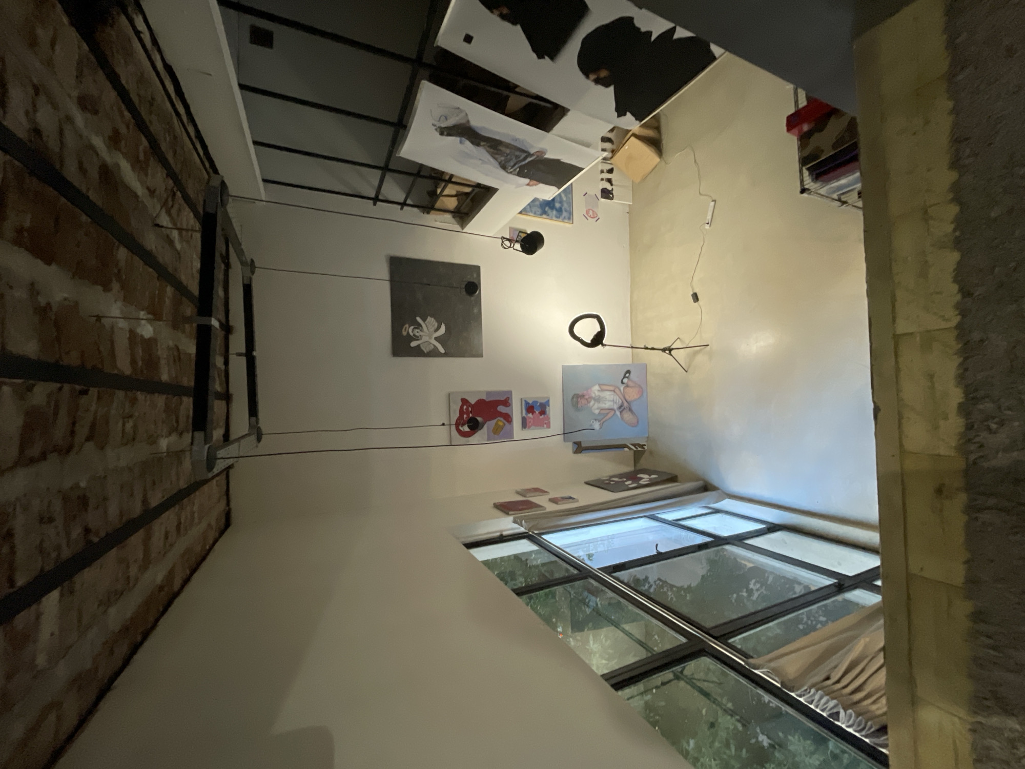 The image depicts an interior space with an industrial aesthetic. The left wall is exposed brick, while the right wall is painted white. The ceiling has exposed black metal beams, and several canvases are suspended from the ceiling, displaying various abstract and figurative images. Additional artwork is displayed on the white wall. A large window with a black frame and multiple panes is visible at the bottom of the frame, offering a view of foliage. There's also a ring light present. The location is likely an art studio or gallery in a building with older construction elements. There is no verifiable information to determine whether this scene is in Buenos Aires, Argentina.