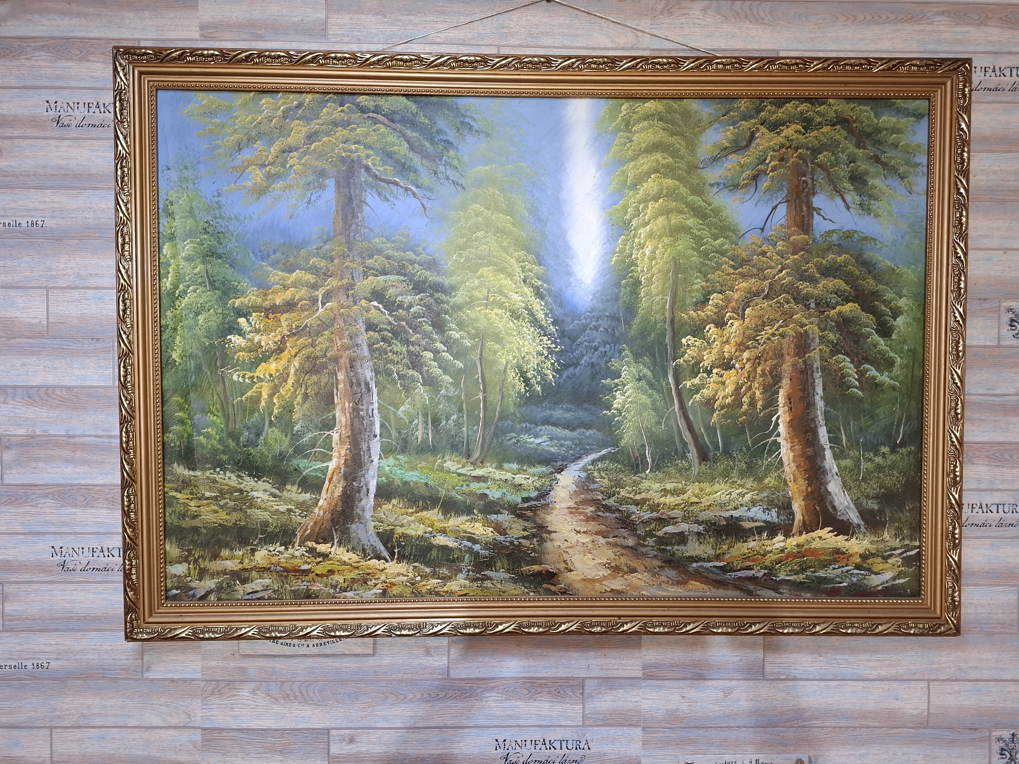The image shows a framed oil painting of a forest scene with a dirt path winding through it. Sunlight streams down from the top center of the painting, illuminating the path and the surrounding trees. The trees are tall and have dense foliage, with some appearing to be evergreens. The forest floor is covered in moss, rocks, and fallen leaves. The painting is hung on a wall with a wood-plank texture. There are watermarks on the wall that appear to be text and logos, some of which are partially visible. The provided crops offer closer views of the painting's frame and texture, as well as some of the text watermarks. The location in Lisakovsk, Kazakhstan is not depicted or implied by the image itself.