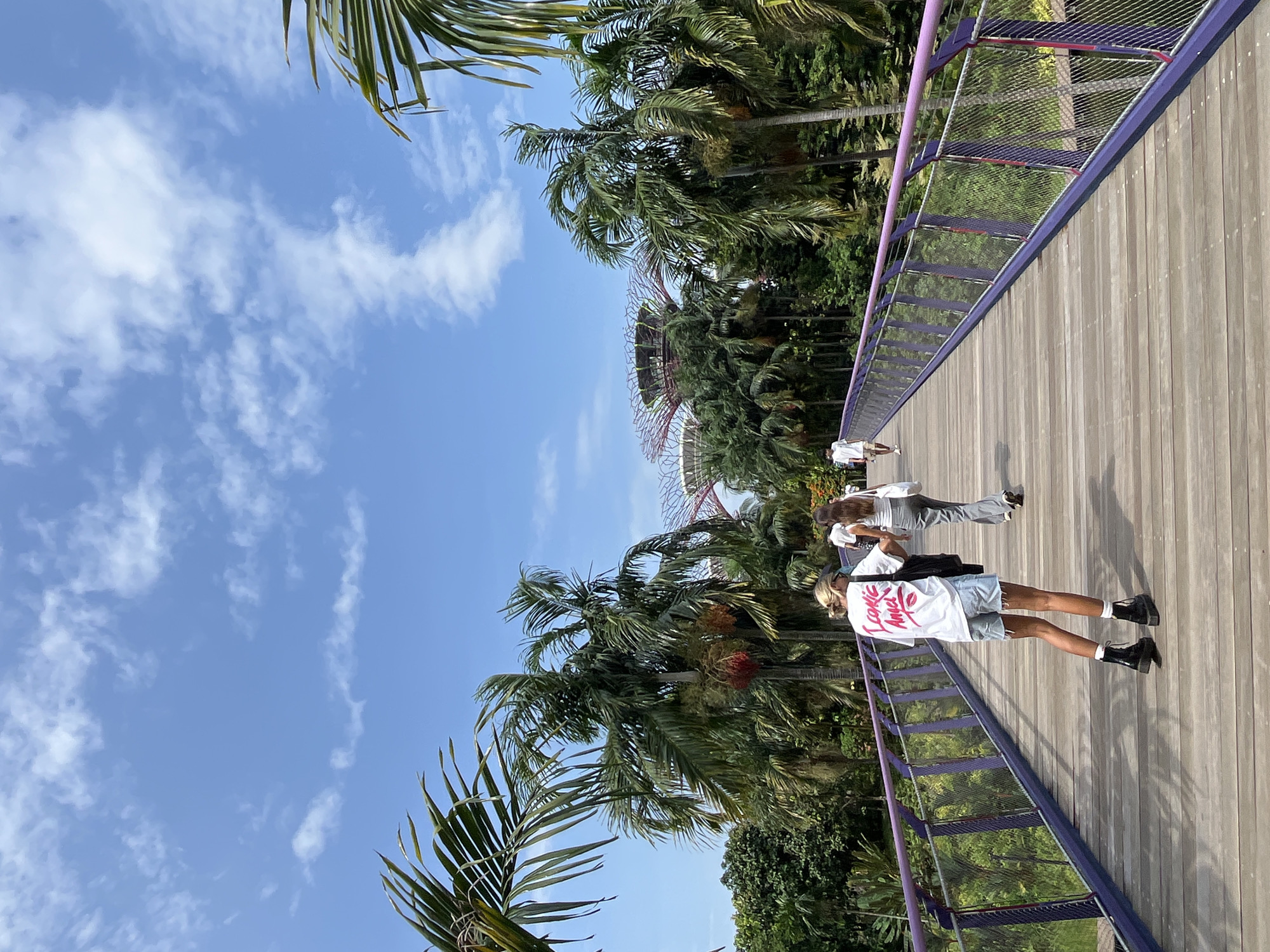 The image shows a view of the OCBC Skyway at Gardens by the Bay, Singapore. In the foreground, a wooden walkway with purple railings and metal mesh fencing leads into the distance. Two women are walking away from the camera on the walkway. The woman in the front is wearing a white t-shirt with a graphic print, denim shorts, and black boots. The woman behind her is wearing a white top and grey pants. In the background, lush green tropical trees and palm trees are visible. The iconic Supertree Grove structures of Gardens by the Bay are partially visible in the distance, along with a clear blue sky with scattered white clouds.