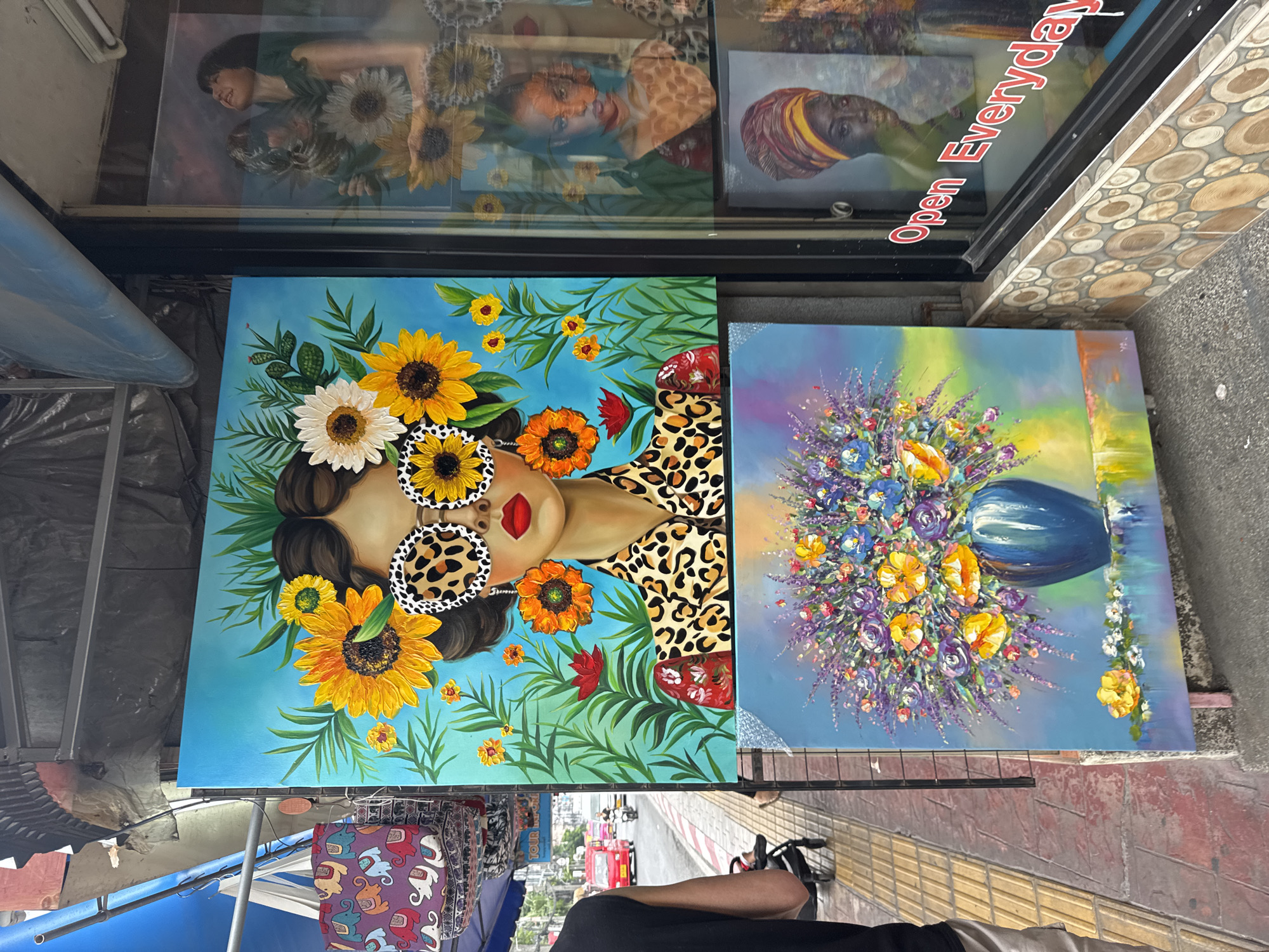 The image shows a street art stall in Patong, Thailand. The stall has two large paintings displayed on the ground: one of a woman with leopard print clothing and sunflower-shaped sunglasses, and another of a vase of flowers. Above these, a glass storefront reflects other paintings, including portraits of women. The storefront also has an "Open Everyday" sign. A person wearing black clothing and white shoes is visible in the foreground, seemingly observing the art. Other stalls and vehicles can be seen in the background, suggesting a busy street scene.