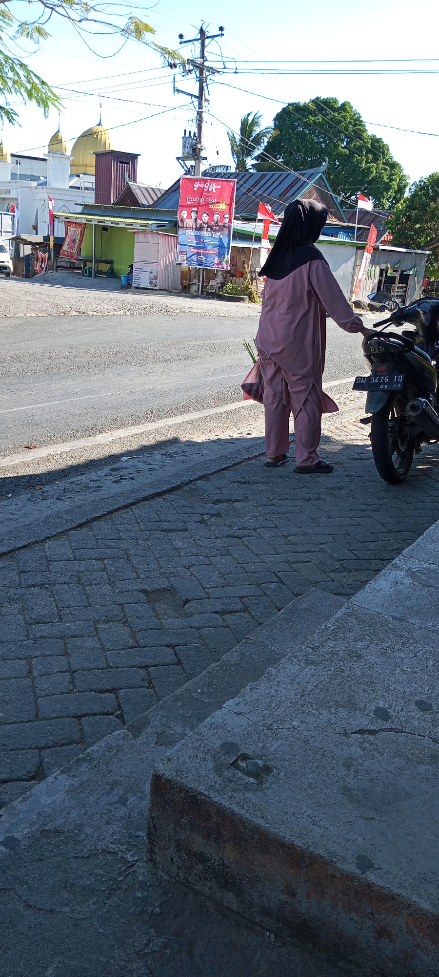A woman wearing a pink abaya and black hijab stands beside a motorcycle on a street in Pakkanna, Indonesia. She is holding a red and white striped plastic bag with green onions protruding from the top. The street is paved with asphalt, and the sidewalk is made of interlocking bricks. In the background, a mosque with a golden dome is visible, along with several small buildings and a utility pole with electrical wires. A banner hangs from the utility pole advertising "Prolog Fest 2023" and "Peterpan". The sky is clear and blue.