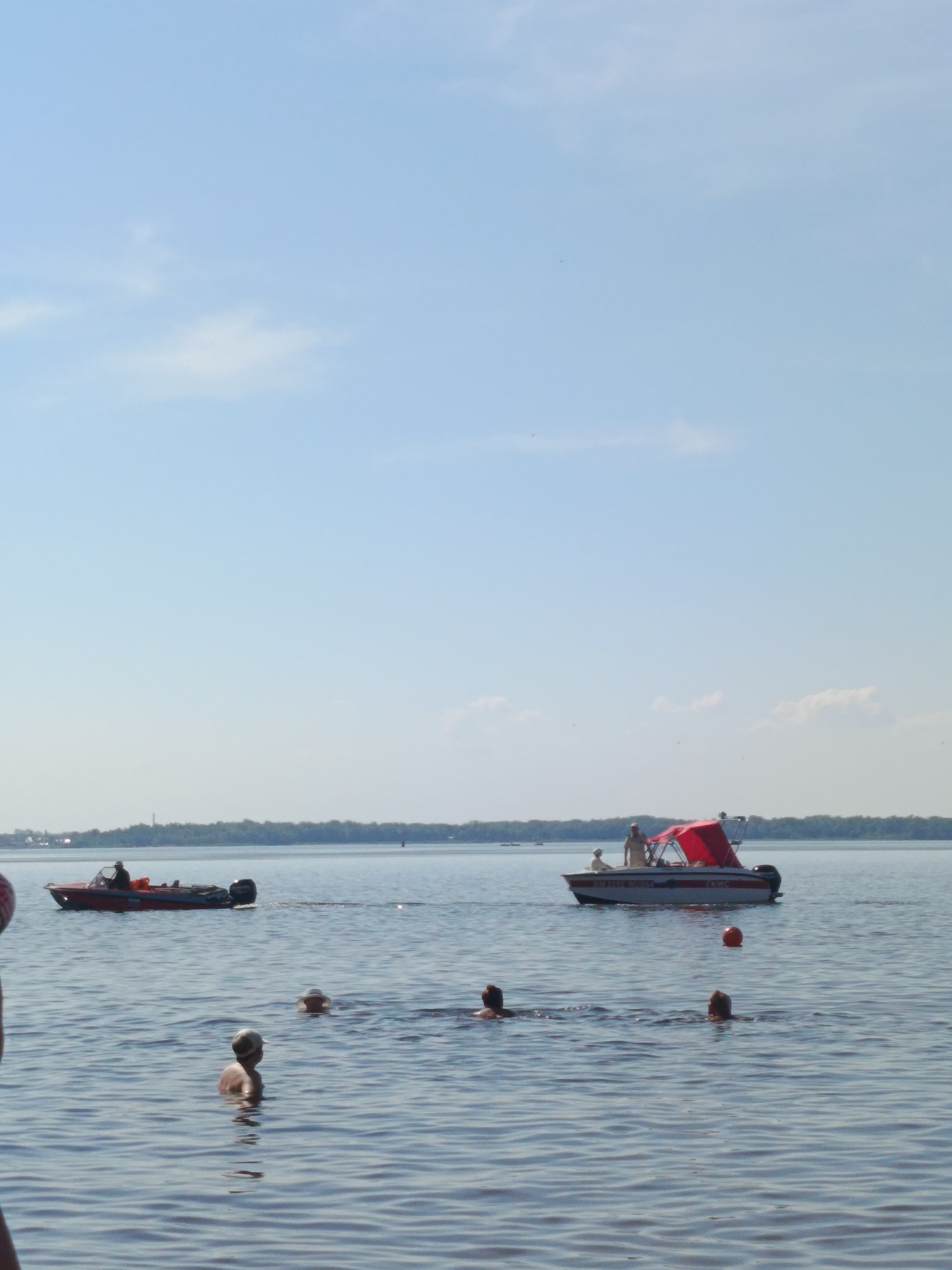 The image depicts a body of water with several individuals and boats. In the foreground, three people are partially submerged in the water, appearing to be swimming. One person, wearing a light-colored cap, is facing away from the camera, while the heads of two others are visible further in the water. In the middle ground, two boats are present. The boat on the left is red and appears to be moving away from the camera. The boat on the right is white with a red canopy and has several people on board, one of whom is standing. A bright orange buoy is visible in the water between the two boats. The background shows a tree line with a hazy sky above. The water is calm with gentle ripples.