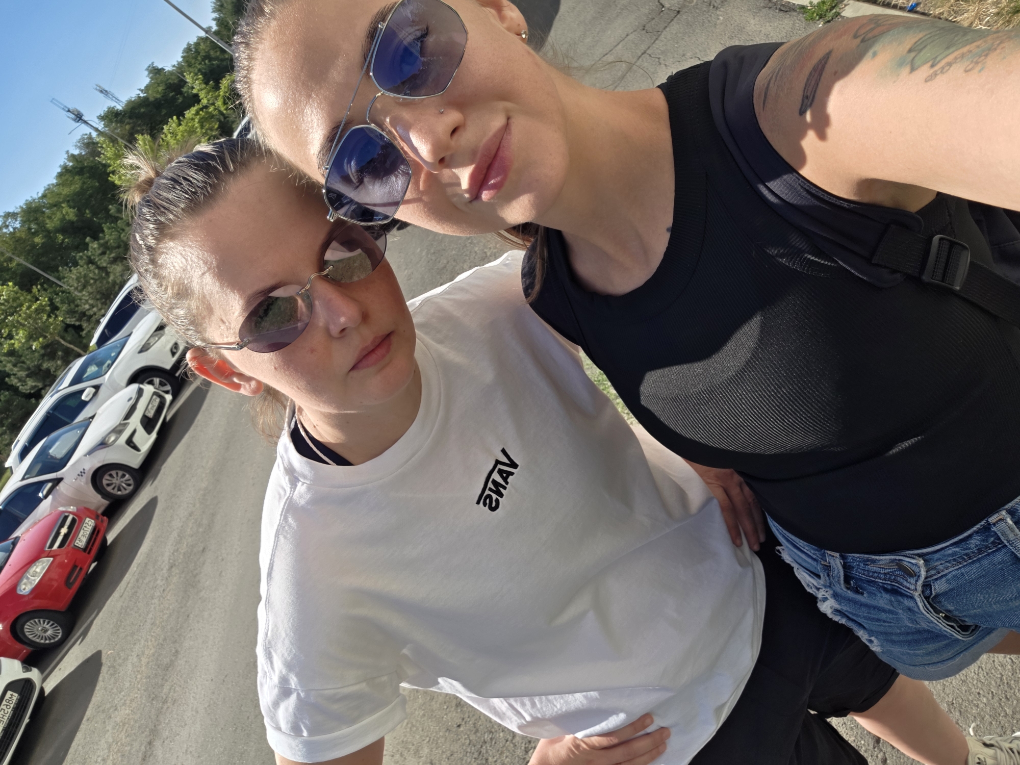 Two individuals are captured in a selfie taken outdoors, likely in a parking lot in Rostov-on-Don, Russia. The background displays a row of parked cars, including a red Chevrolet and several white vehicles. The individuals are wearing sunglasses, with one sporting a white t-shirt bearing the Vans logo and the other a black ribbed tank top. Both appear to be posing for the camera, with their faces angled towards it. The environment suggests a sunny day, with trees visible in the background. The overall scene is casual and appears to be a snapshot of a shared moment.