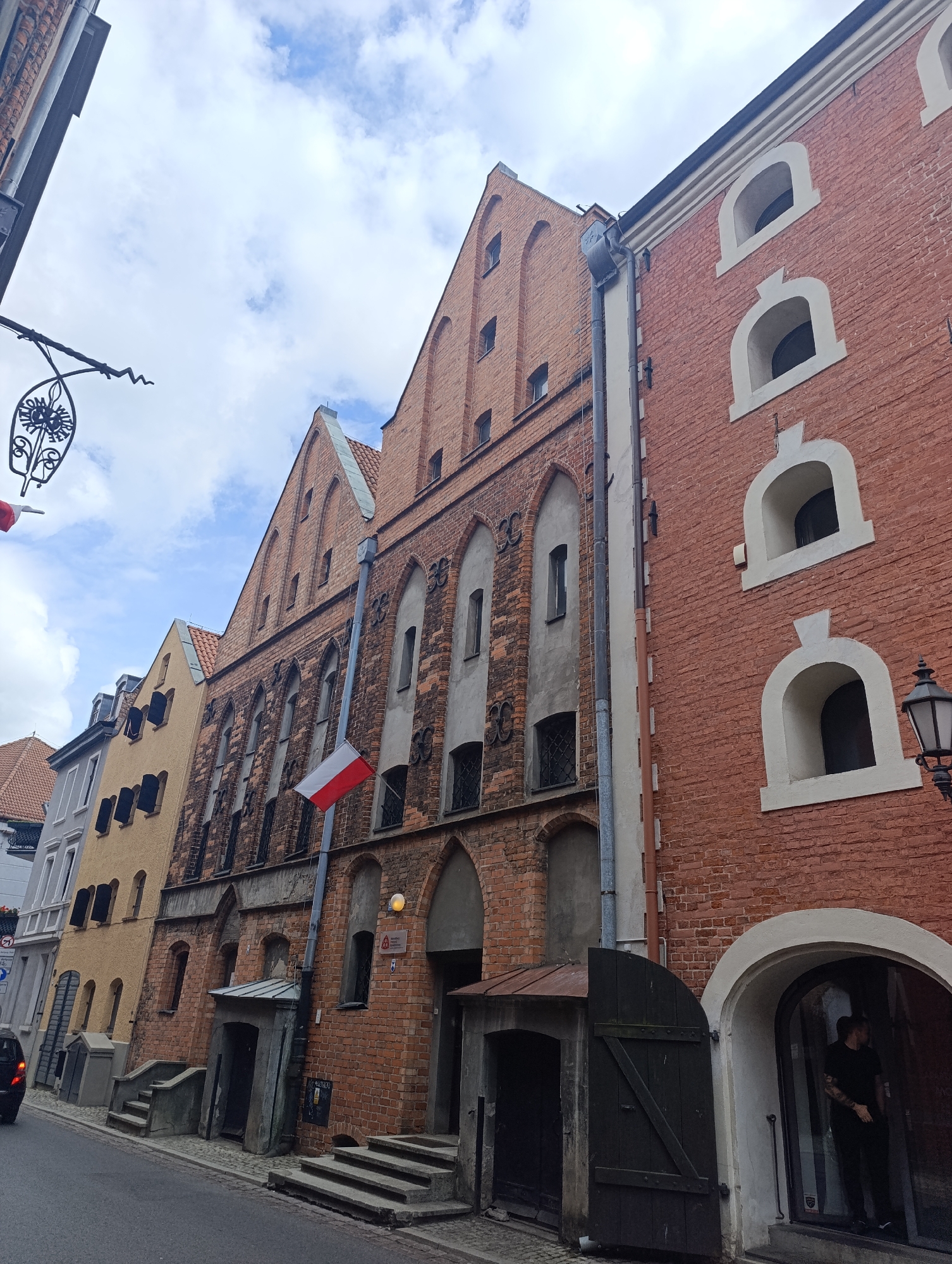 The image depicts a street view in Toruń, Poland, focusing on the facades of historic brick buildings. The sky is partly cloudy, and the day appears to be overcast.

On the right, a red brick building with arched windows, outlined in white stucco, dominates the foreground. Its facade has a rough texture, and a vintage black lantern is mounted on the wall to the right. An arched entryway leads into a dark interior where a person wearing a black t-shirt and pants is visible standing. To the left of this building, another brick building with a steeply pitched gable roof features narrow, Gothic-style windows. A Polish flag is attached to a pole and extends from this building.

Further to the left, a narrower building painted a muted yellow has several dark shutters. Adjacent to it is a lighter-colored building with a grey facade. The street in the foreground is paved with cobblestones, and a grey sidewalk runs along the buildings. A few steps lead up to some of the building entrances.

A black car is partially visible on the left edge of the image, moving away from the camera. A decorative wrought-iron sign hangs from a pole over the street, with a circular emblem featuring floral motifs. The overall impression is of a narrow street lined with old, characteristic European architecture.