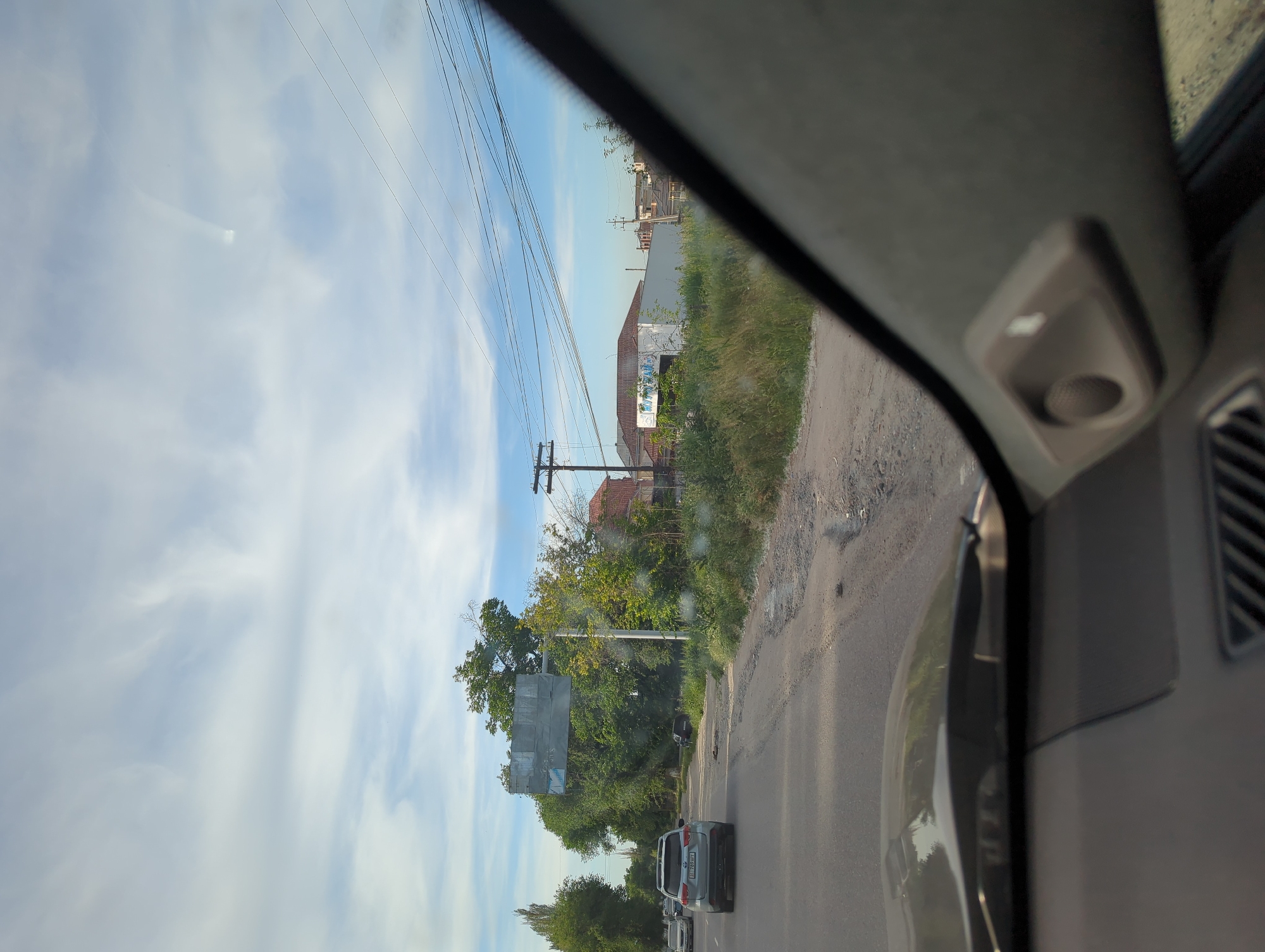 The image depicts a view from inside a car traveling on a paved road in Beş-Küŋgöy, Kyrgyzstan. The sky is visible through the windshield with power lines running across it. To the right of the road is overgrown foliage and dilapidated buildings. There are vehicles in front of the car. The interior of the car, including the interior trim and air vent, is in the foreground.