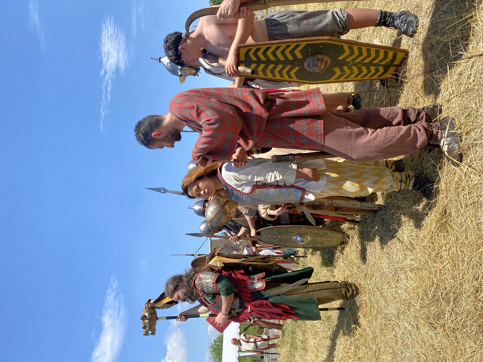 The image captures an outdoor historical reenactment scene in Gigen, Bulgaria, under a clear blue sky with sparse clouds. The ground is covered in dry, mown hay or straw, and strong sunlight creates defined shadows.
In the left foreground, a man with a long brown beard and hair wears a green tunic, a red fringed sash, and scale armor with metallic shoulder pieces. He holds a tall standard topped with a golden animal-headed finial (possibly a draco) on a black and yellow striped pole in his left hand, and a red rectangular object (possibly a book or tablet) in his right. A sword is sheathed at his left hip. His gaze is directed towards the right.
Central to the frame, a person with reddish-brown hair, appearing female, stands wearing a white tunic, scale armor, and yellow-and-white patterned trousers. She faces right, slightly to the front. Behind her, a man with short dark hair and a beard, dressed in a red and blue plaid tunic and dark reddish-brown trousers, leans forward, his hands near her right shoulder, appearing to adjust her attire or instruct her. His head is tilted downwards, focused on her.
To the far right, a young man, shirtless, wears grey trousers and black boots over patterned socks. He holds an elongated, scutum-like shield with a green field and yellow chevron pattern in his left hand. In his right hand, he grasps a large, curved metal implement with a wooden handle; an additional hand is also visible assisting in holding this tool. He looks towards the left, observing the central interaction.
In the background, several other reenactors are partially visible, equipped with spears, metal helmets (galeas), and various shields, forming a line or group. A white tent is discernible in the far left background, and distant greenery marks the horizon.