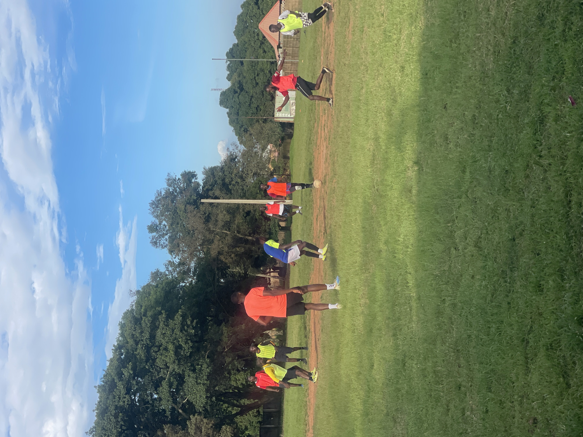 In Jos, Nigeria, a group of young men is engaged in a soccer game on a dusty field surrounded by lush green grass and trees. The sky above is a vibrant blue with scattered white clouds. The players are dressed in casual athletic wear, some in brightly colored t-shirts and shorts, others in bibs, indicating team affiliations. The game appears to be in progress, with some players actively chasing a soccer ball, while others are positioned strategically on the field. The overall atmosphere suggests a spirited and energetic sports activity taking place outdoors.