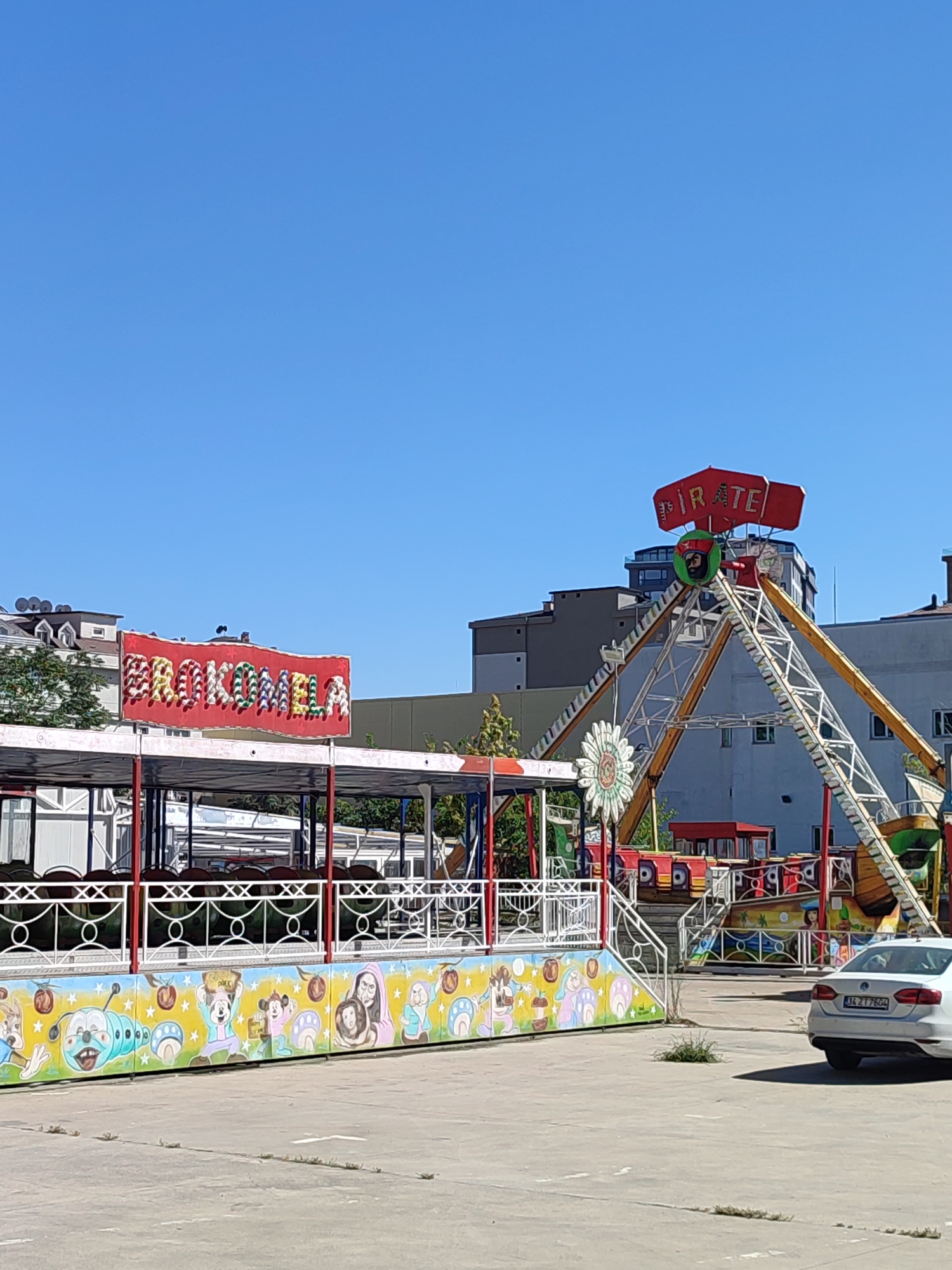 In Maltepe, Turkey, on a clear, sunny day, an outdoor amusement park is depicted. The foreground is dominated by a low-lying amusement ride with a colorful mural depicting cartoon characters such as Mickey Mouse and a caterpillar. A white railing with red posts encloses this ride. To the left, a red banner with illuminated letters that spell "ERÖKKOMELA" is prominently displayed above the ride.
In the background, a larger amusement ride, a pirate ship, stands tall. Its structure is triangular with yellow support beams, and a red sign at the top reads "PIRATE." A circular depiction of a pirate's face is visible on the ride's structure. Various colorful cars of the pirate ship ride are visible at the base of its structure. Behind the rides, several multi-story buildings are visible, indicating an urban setting. A white car with license plate "34 ZTT 7604" is parked to the right of the image. The ground is a paved, gray surface, with patches of dry grass scattered throughout. The sky is a clear, vibrant blue.