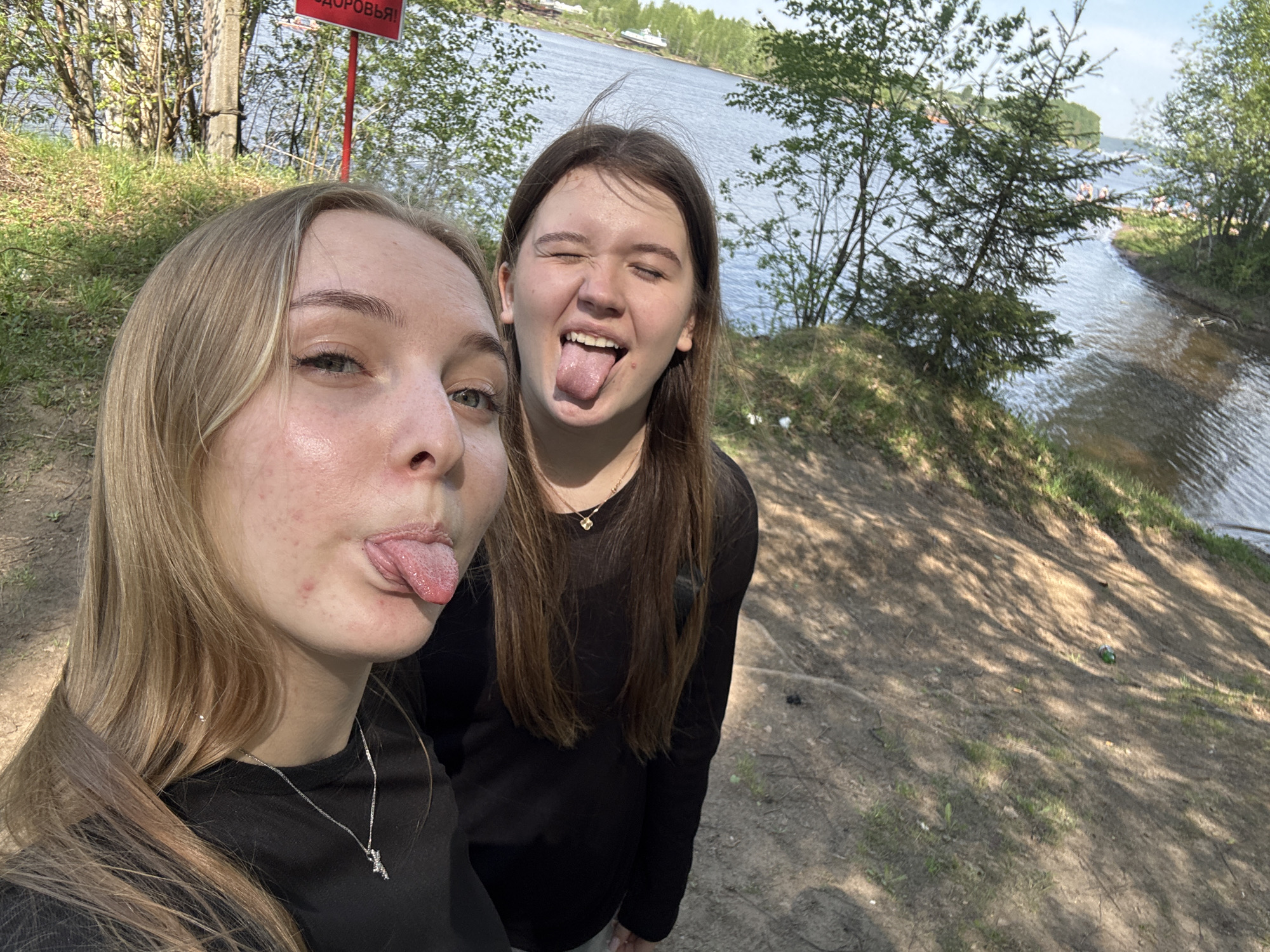 Two young women stand on a grassy, sandy riverbank. The woman on the left has blonde hair and is wearing a black long-sleeved shirt with a silver pendant necklace. The woman on the right has brown hair and is wearing a black long-sleeved shirt with a gold pendant necklace. Both women are sticking out their tongues and posing for the camera. The background includes trees, grass, and a river. A red sign with Cyrillic script is visible. The location is Perm, Russia, on the bank of the Kama River.